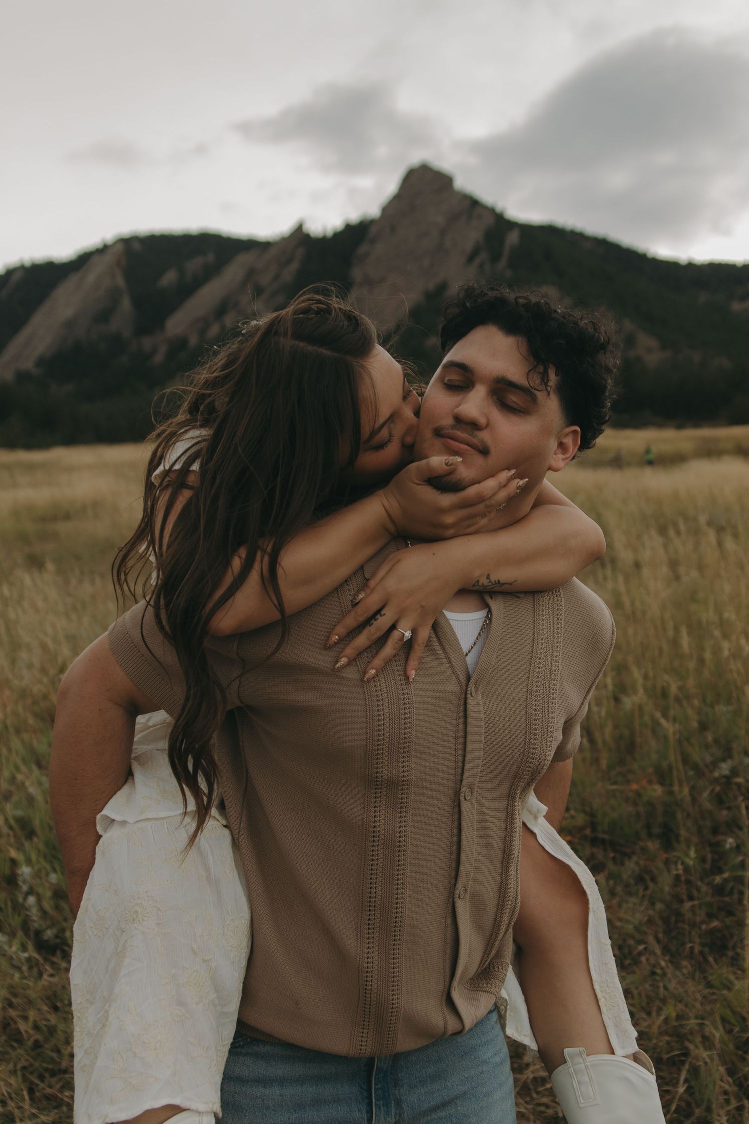 Couple shares a tender piggyback kiss in a golden field with the Flatirons mountains in the background.