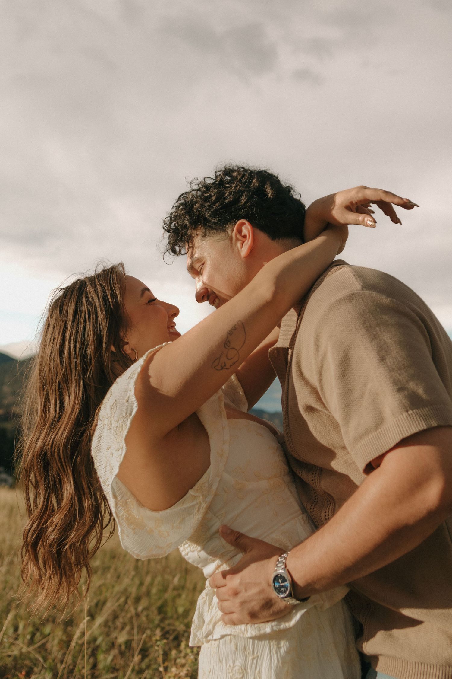 Couple embracing and almost kissing in a golden field with mountains in the background.