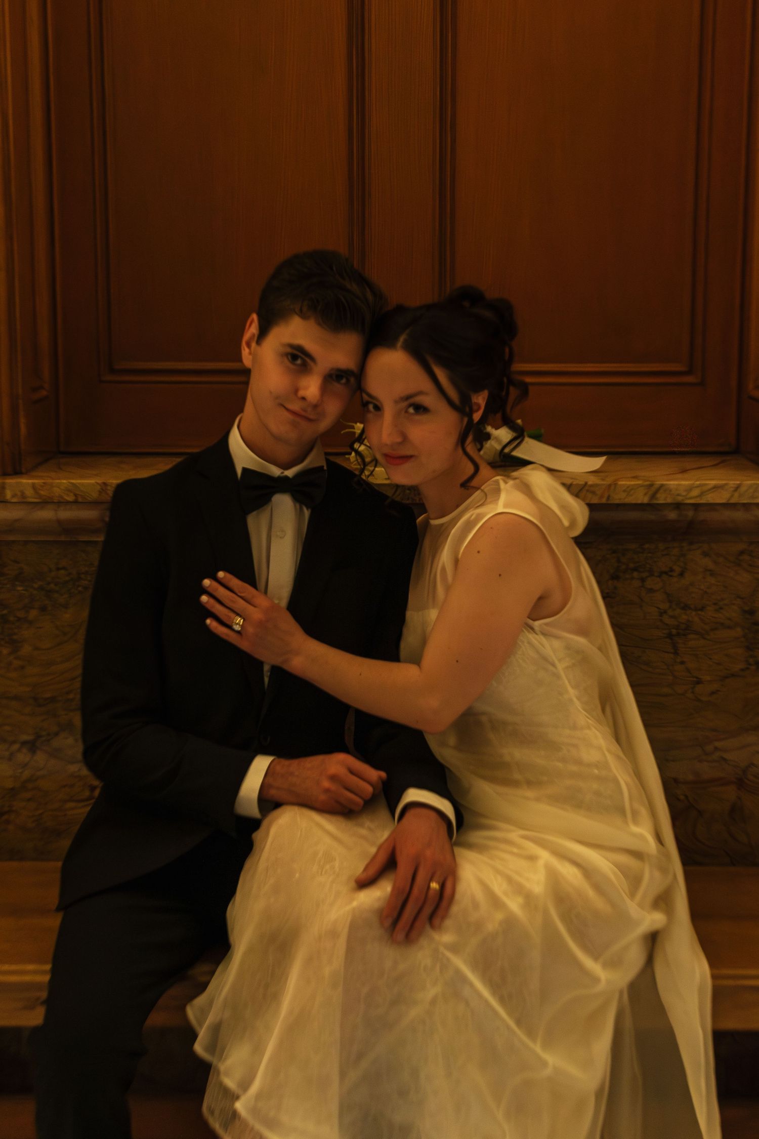 A couple in formal wedding attire sits intimately on an elegant bench.