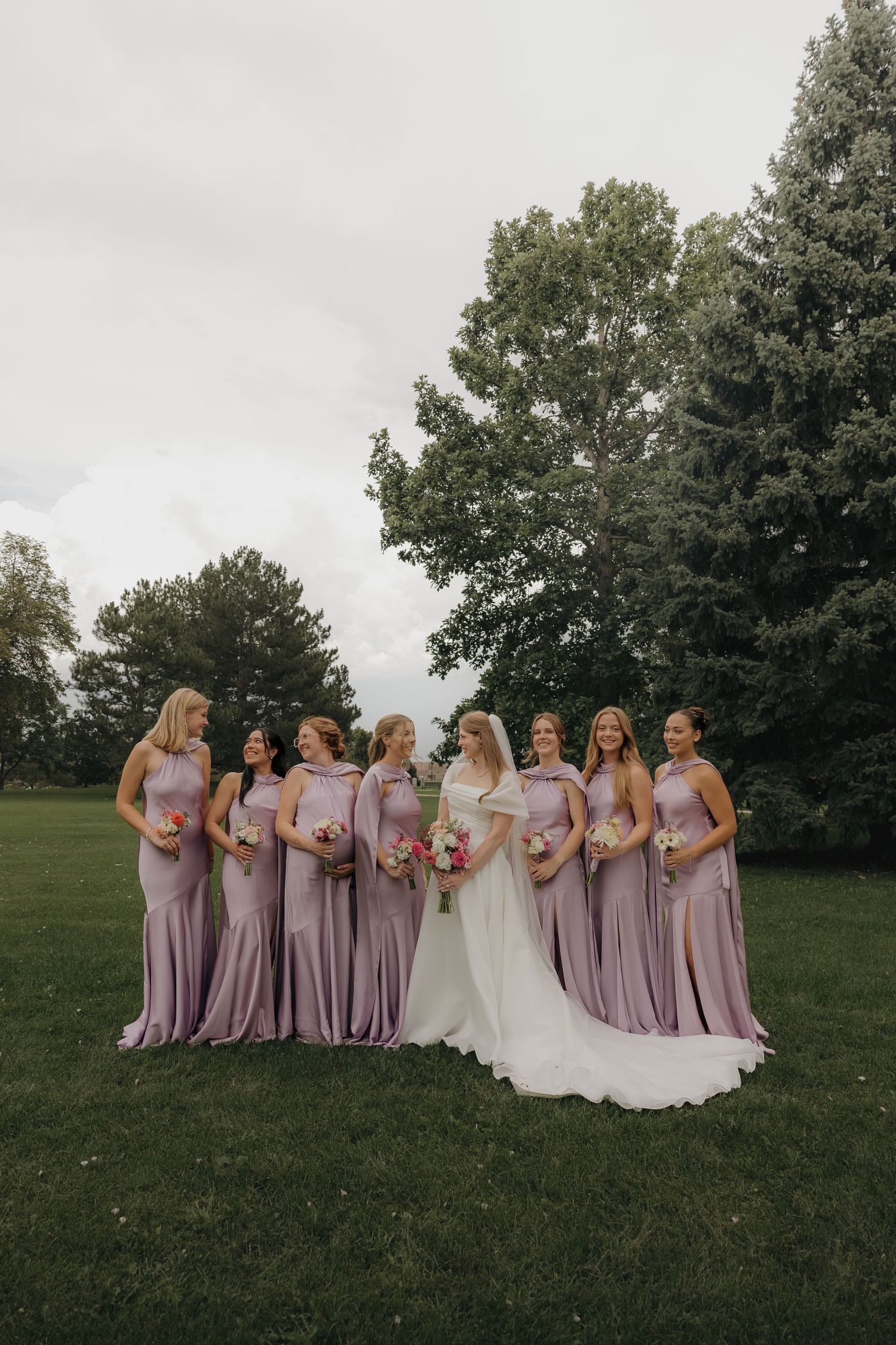 Bride in white gown posing with bridesmaids in lavender dresses on green lawn.
