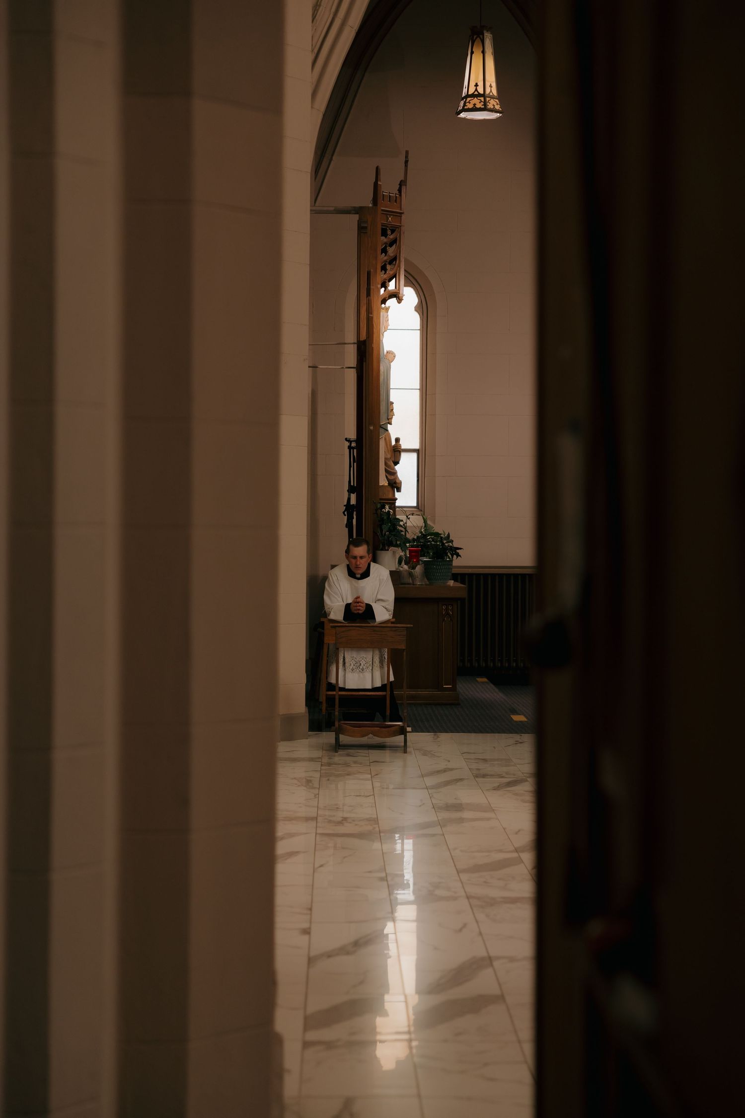 Solitary figure seated in prayer in a dimly lit church corridor with marble floors.