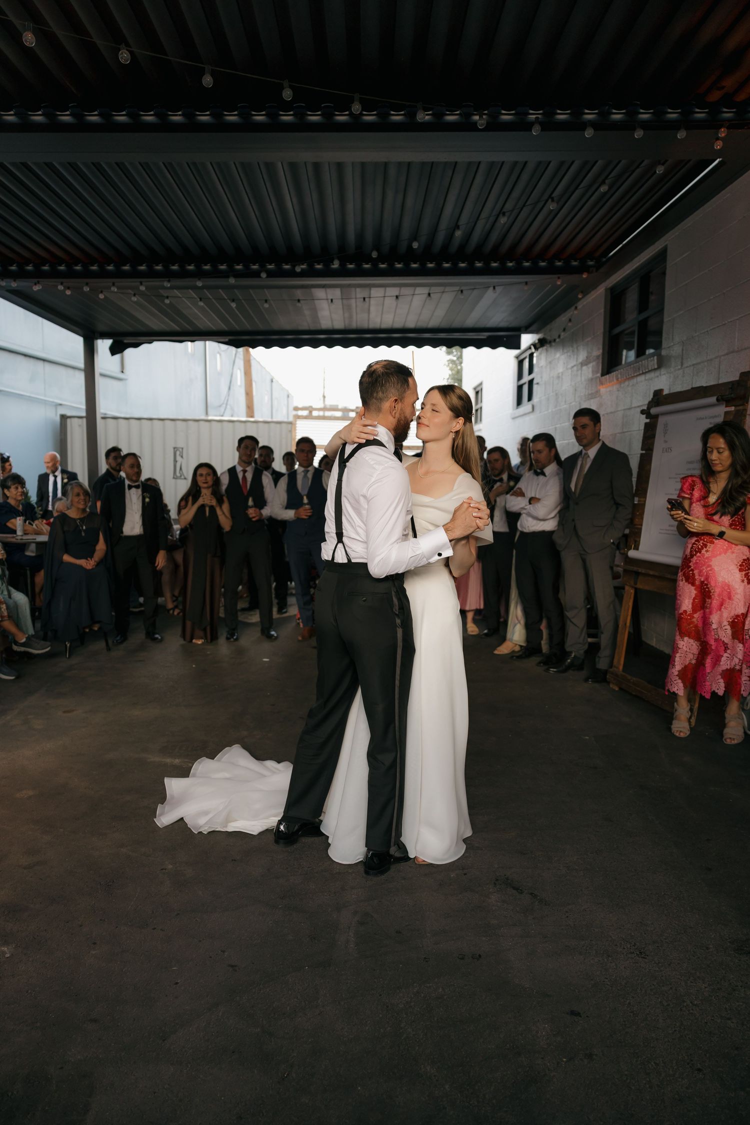 Bride and groom sharing romantic first dance under industrial ceiling while wedding guests watch and photograph.