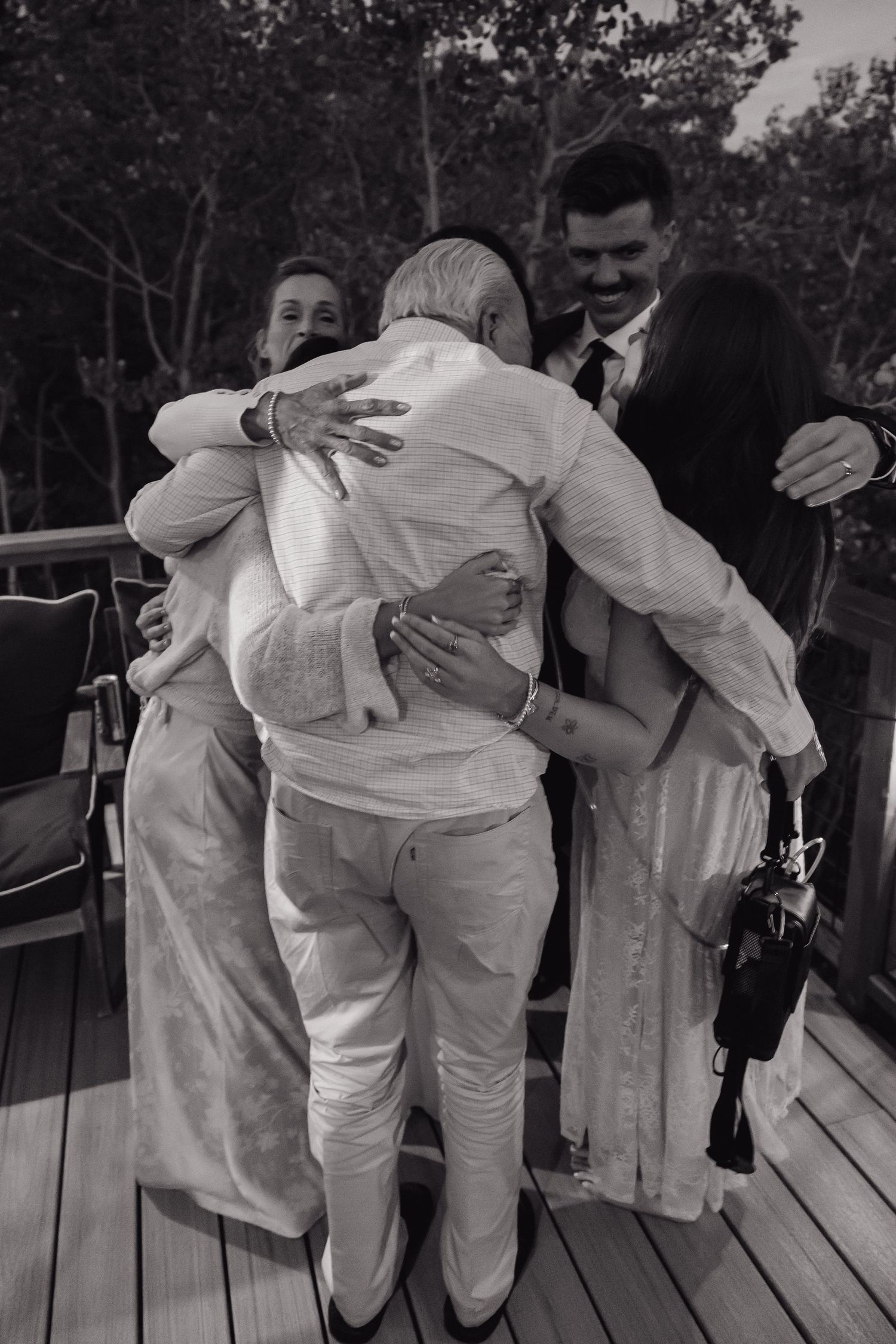 Black and white photo of wedding party sharing a warm group hug on a wooden deck surrounded by trees.