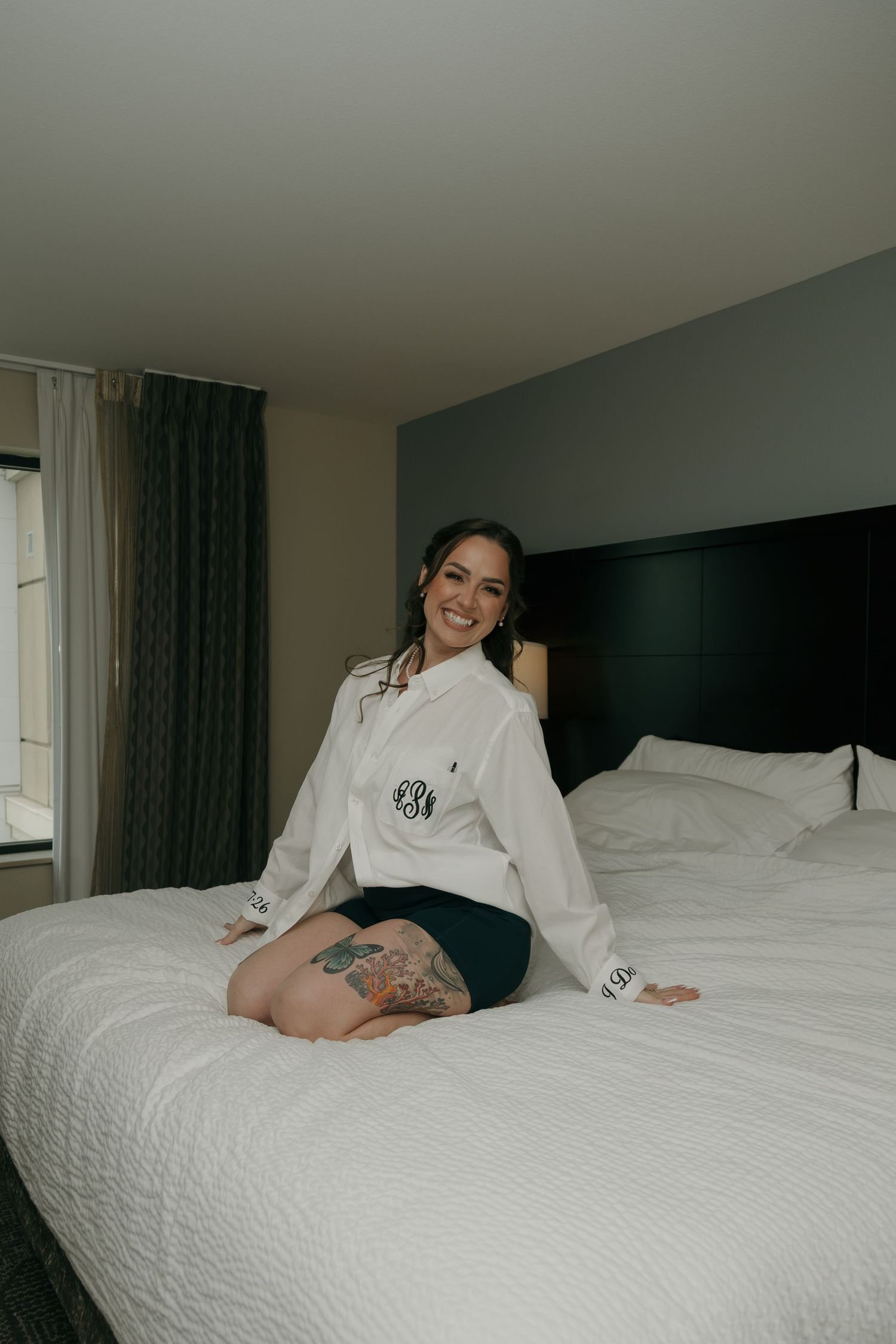 Smiling bride in monogrammed white shirt sitting on hotel bed before wedding day preparations.