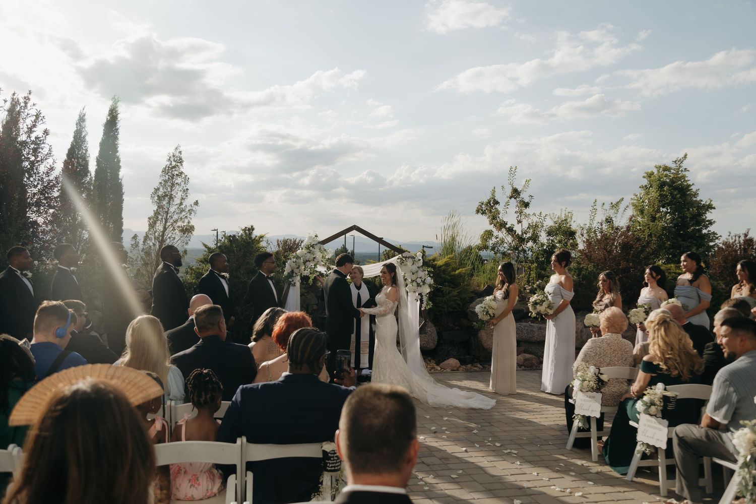 Wide shot of outdoor wedding ceremony with guests seated as couple exchanges vows at floral altar.