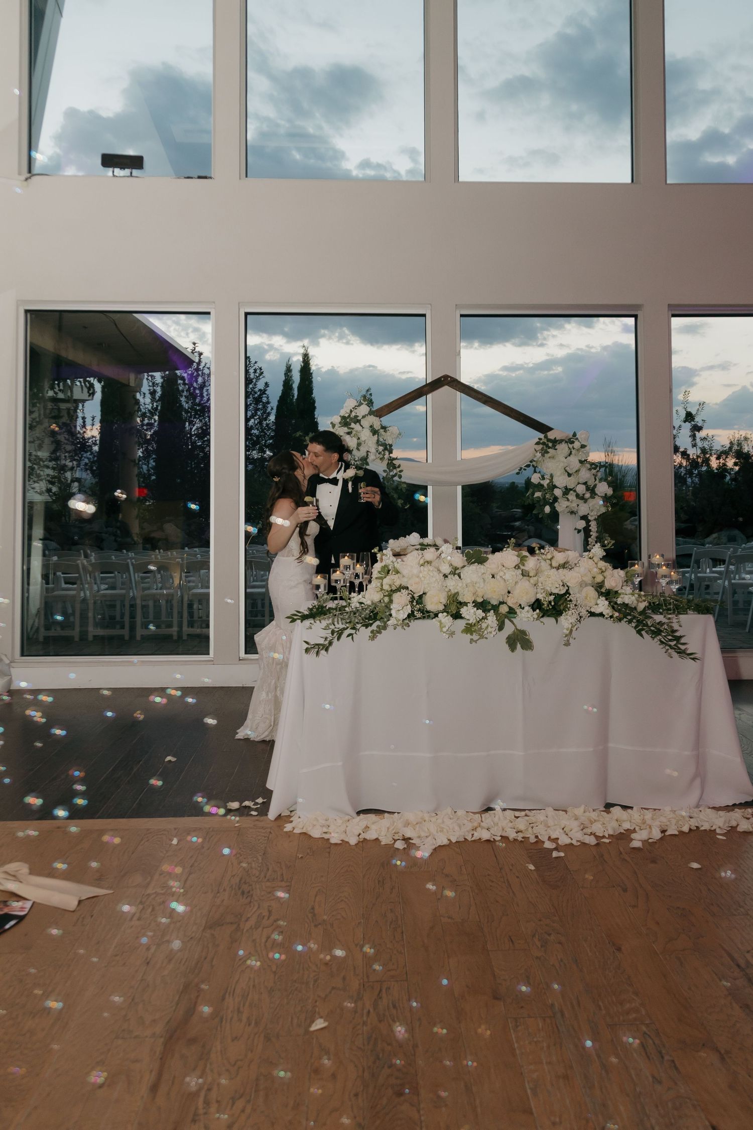 Newlyweds kissing at sweetheart table adorned with white florals during wedding reception.