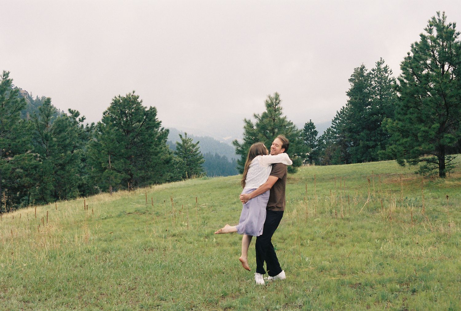 Man lifting woman in embrace on green meadow with pine trees behind.