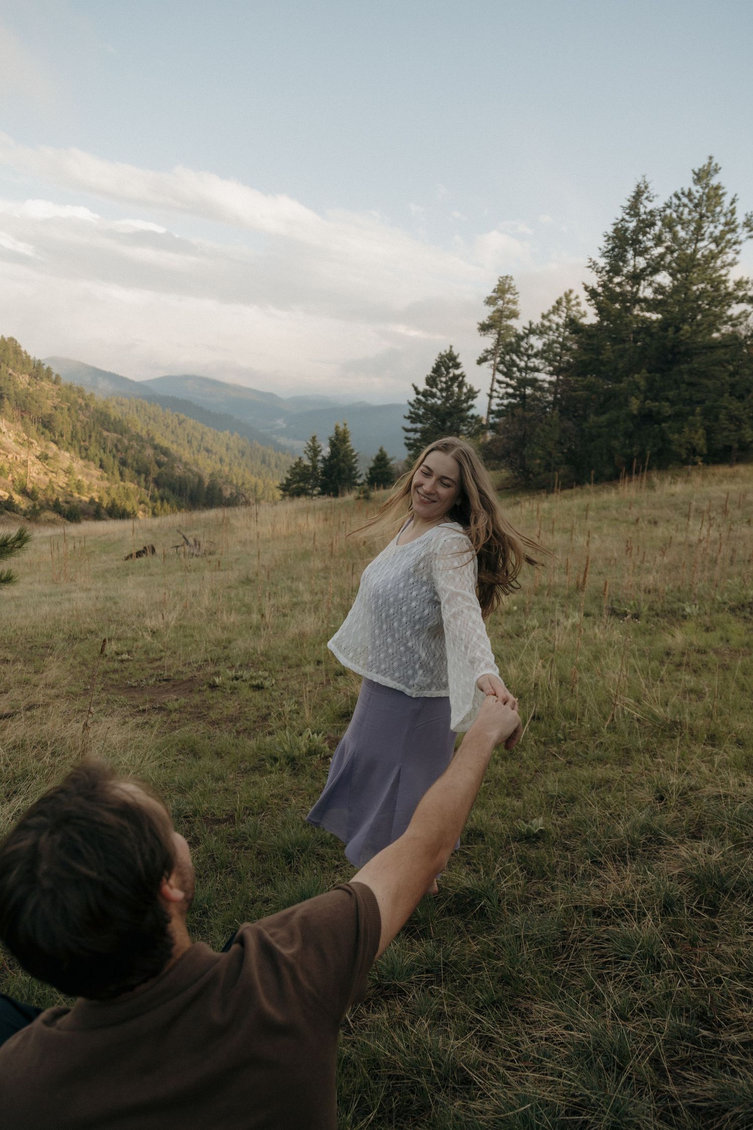 A couple holds hands in a mountain meadow, woman smiling and twirling in a lace top and gray skirt at dusk.