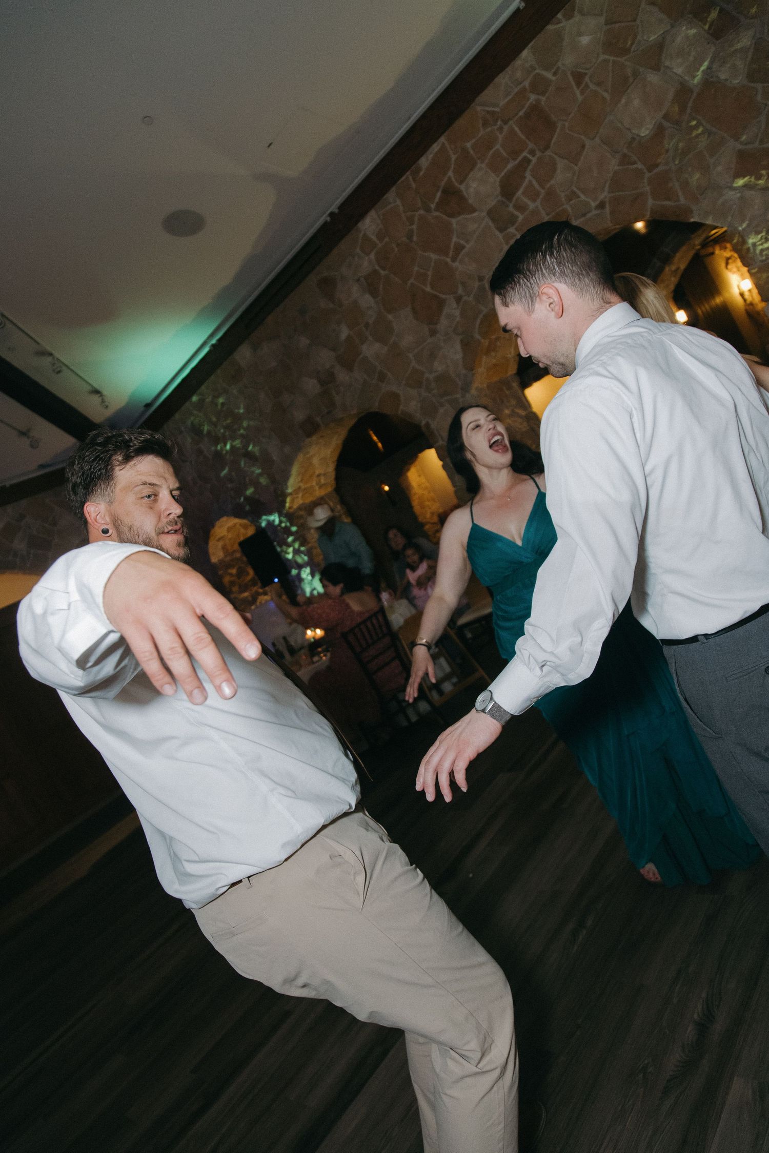 Two men and a woman laugh and dance together at a lively wedding reception.