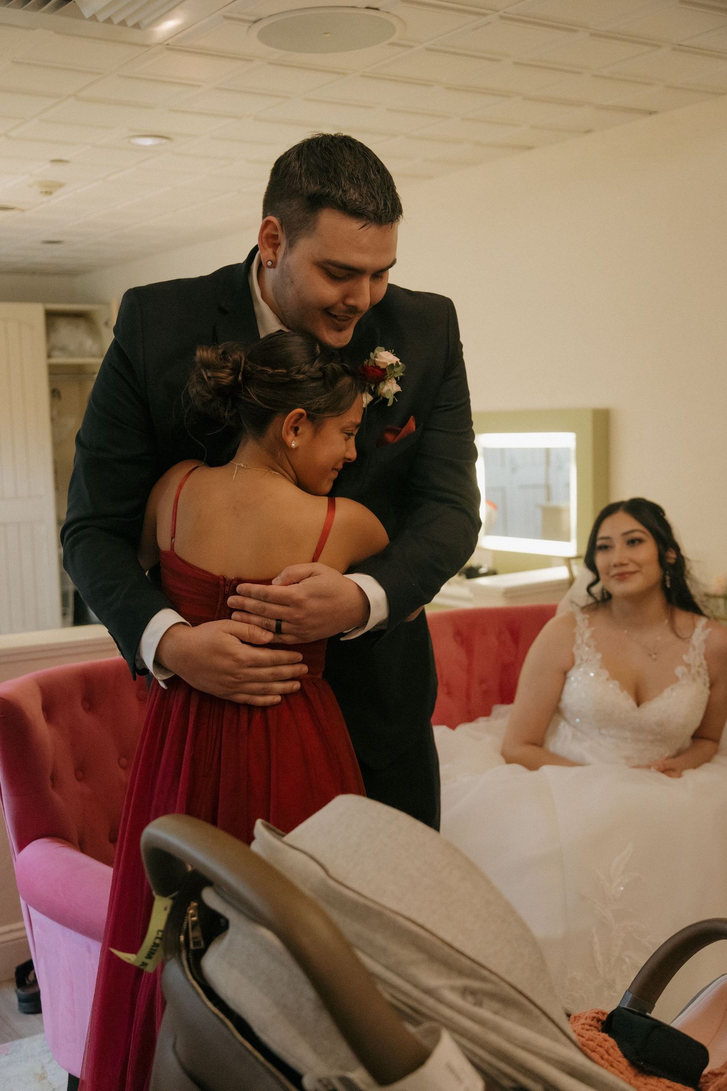 Groom in black suit hugs bridesmaid in red dress while bride watches in bridal suite.