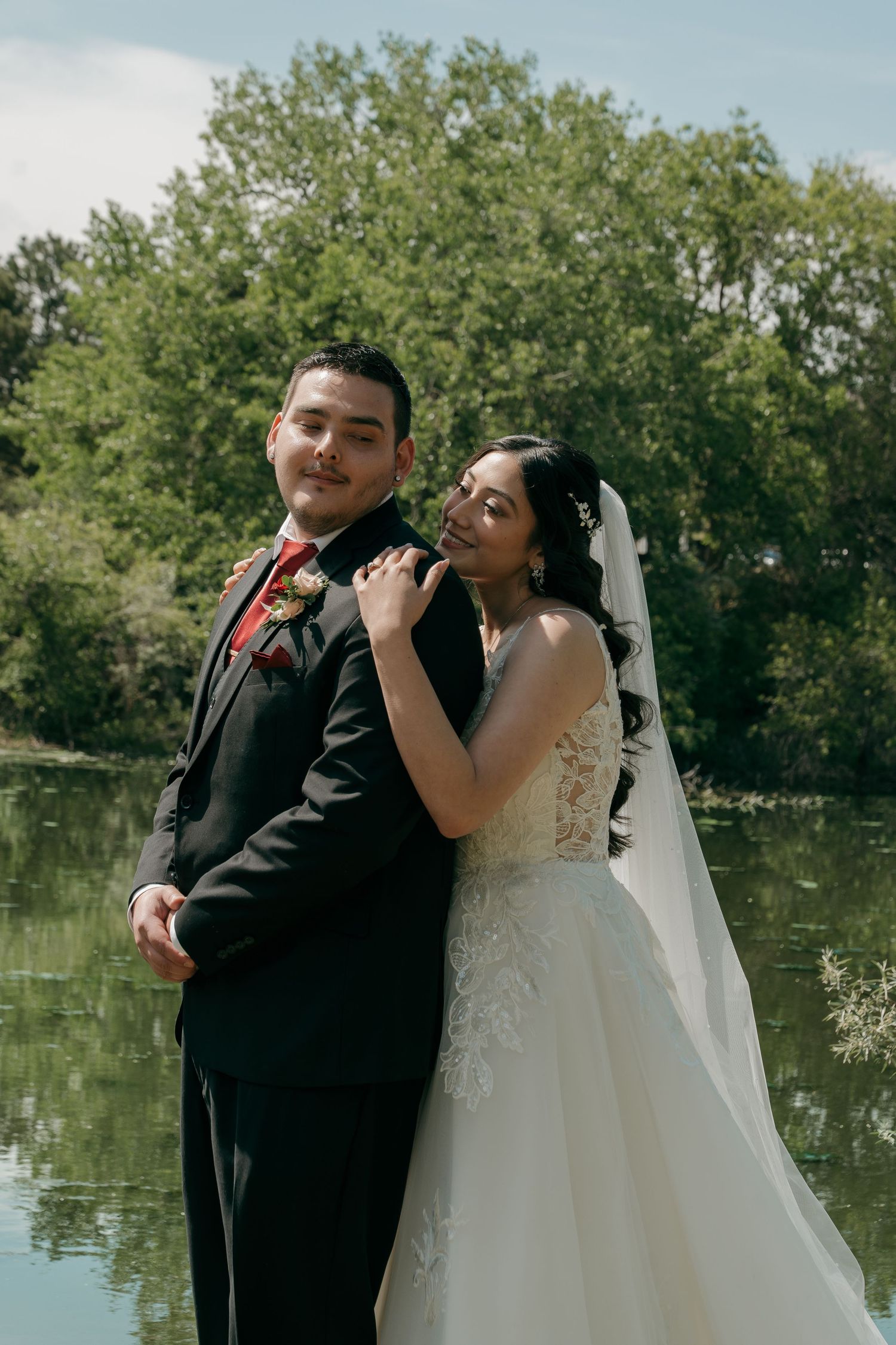 Bride in lace wedding gown embracing groom in black tuxedo near a reflective lake surrounded by trees.
