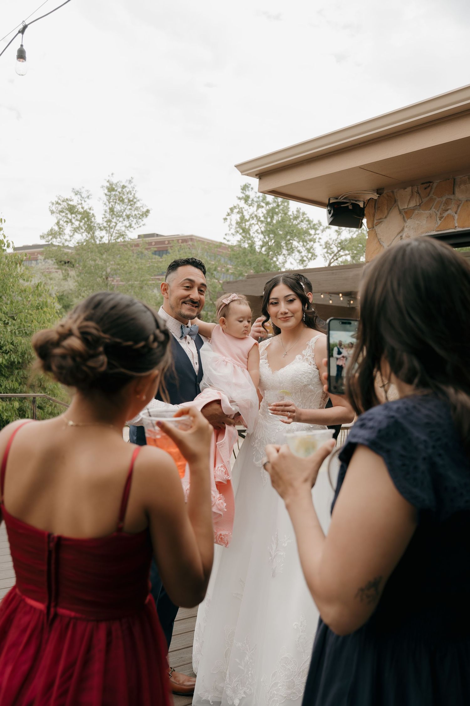 Bride and groom holding baby girl in pink dress greet guests at outdoor wedding reception.