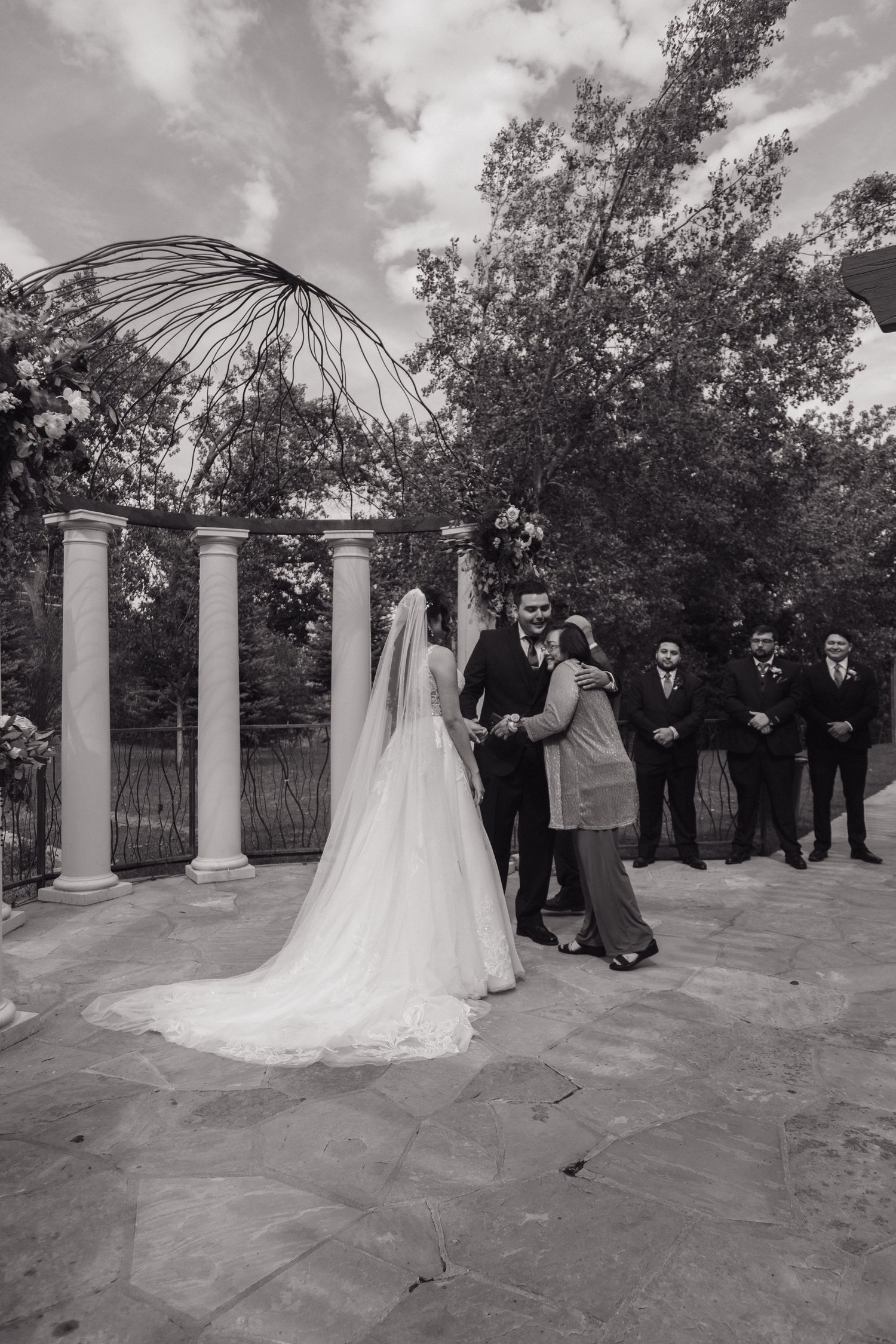 Black and white photo of bride and groom exchanging vows at outdoor ceremony with columns and groomsmen.
