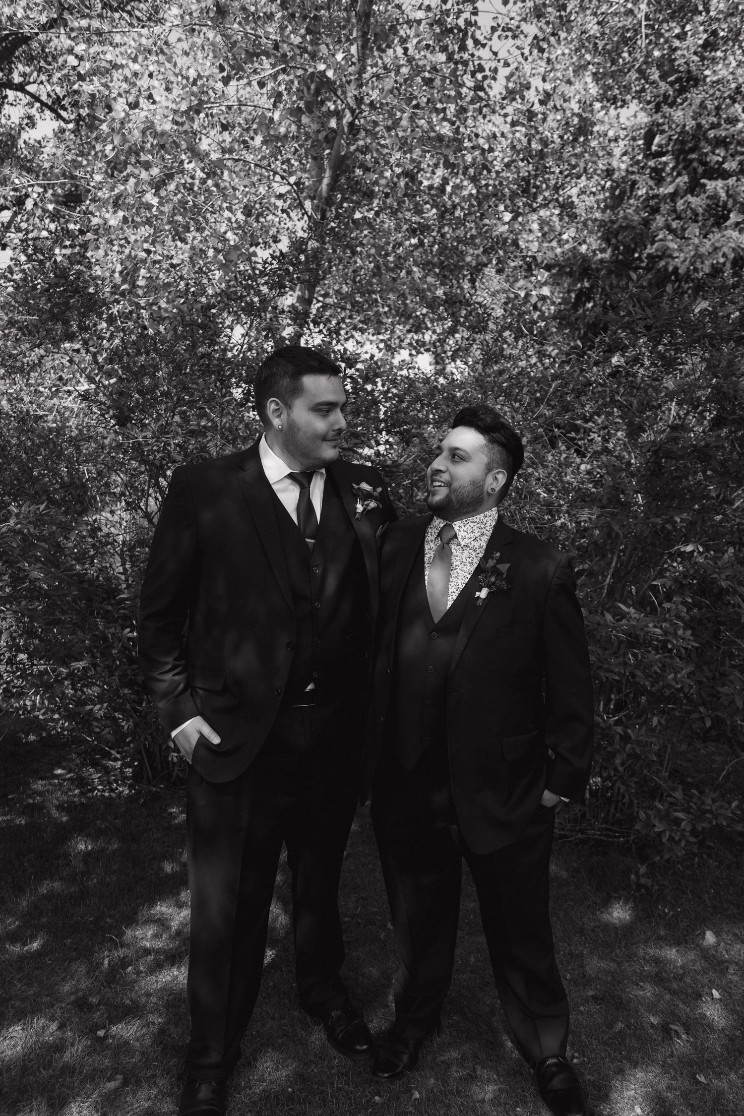Black and white photo of two grooms in suits sharing a moment outdoors surrounded by trees on their wedding day.