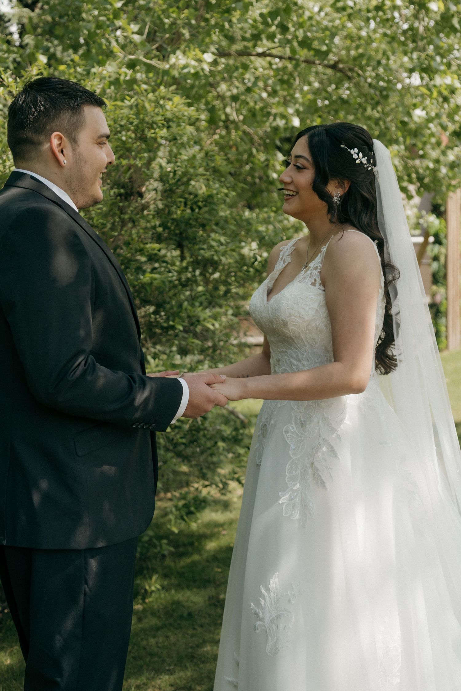 Smiling bride in lace gown and veil holding hands with groom in black suit outdoors.