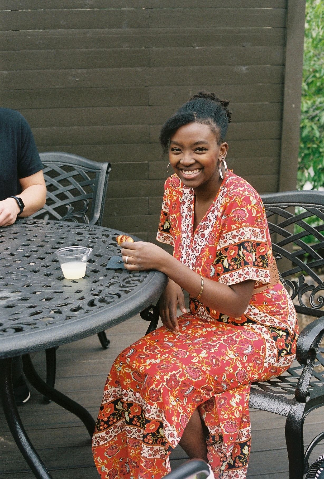 Smiling woman in red floral dress sitting at ornate patio table at outdoor wedding reception.