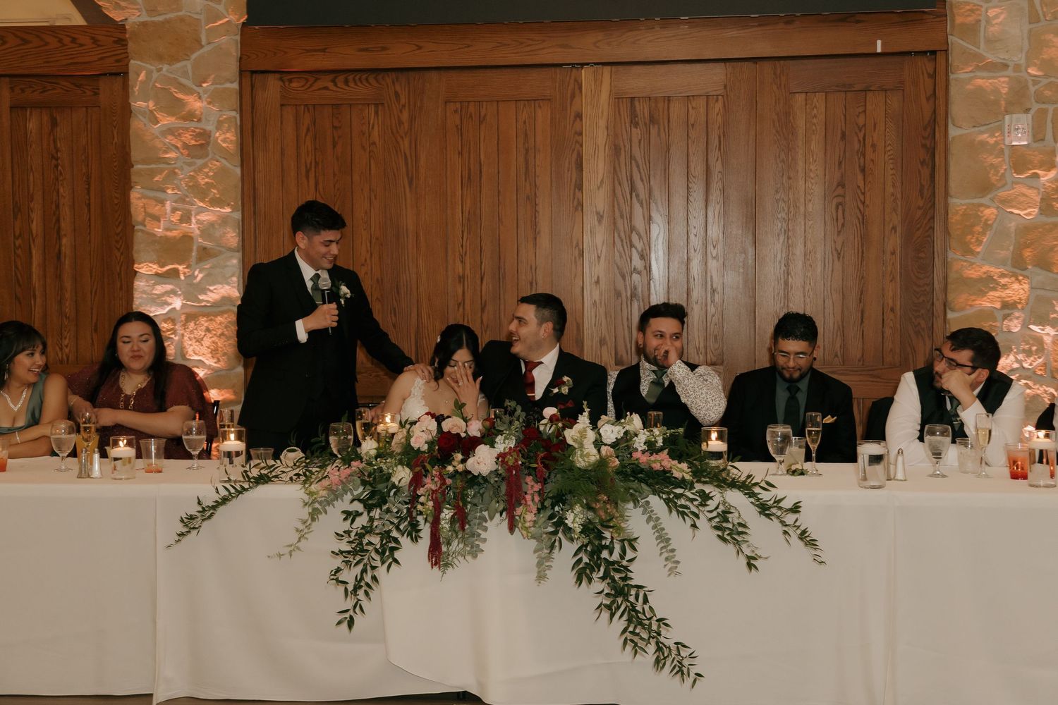 Best man delivering speech at wedding reception while bride and groom smile at decorated head table.