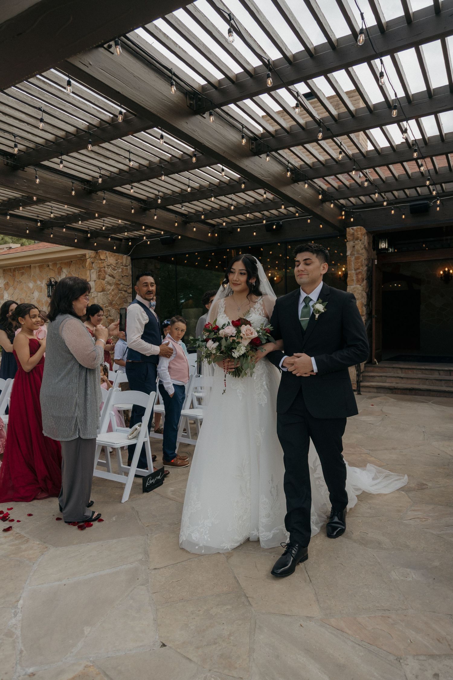 Newlyweds walk down aisle under string lights as guests celebrate at outdoor wedding ceremony.