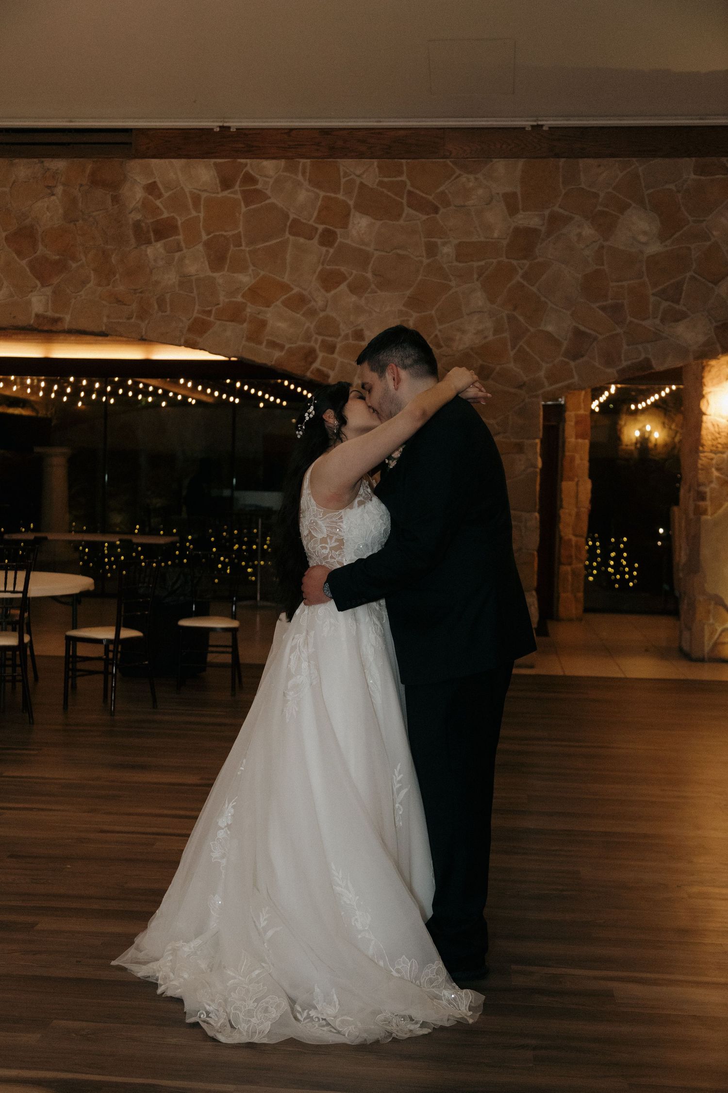 Newlyweds share intimate first dance embrace on empty dance floor in warmly lit stone reception hall.