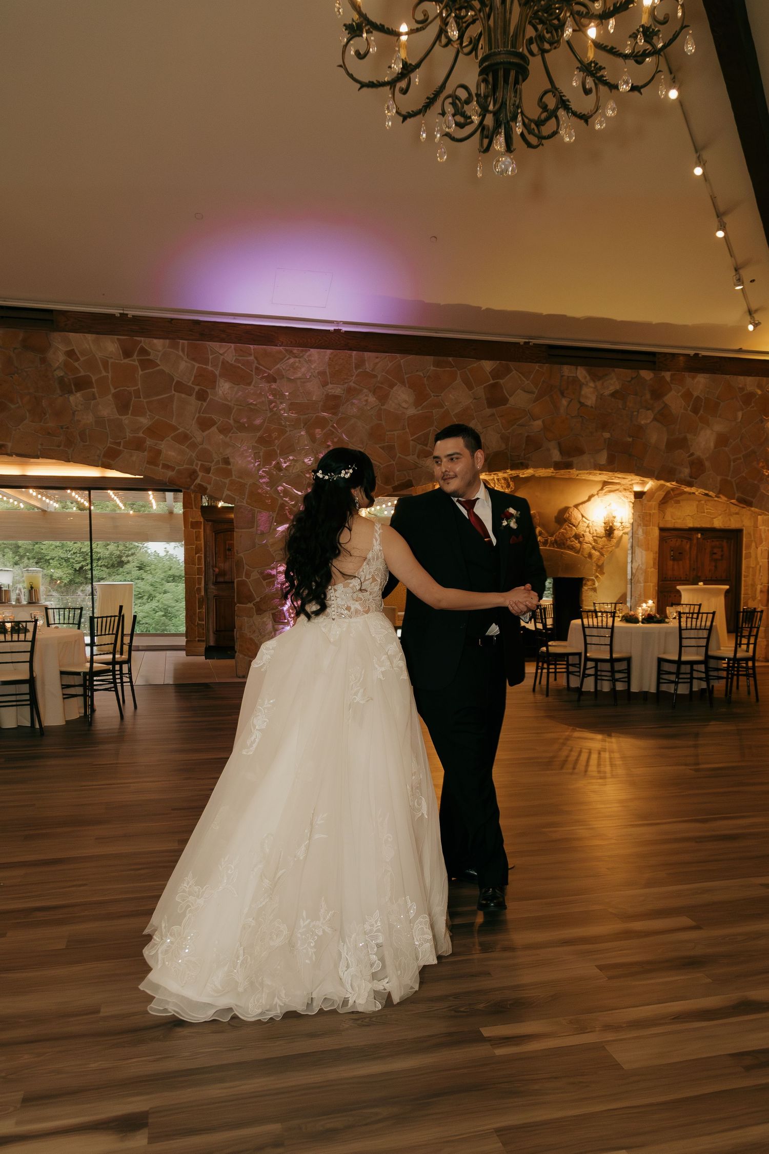 Bride and groom share first dance under chandelier in rustic stone wedding reception venue.