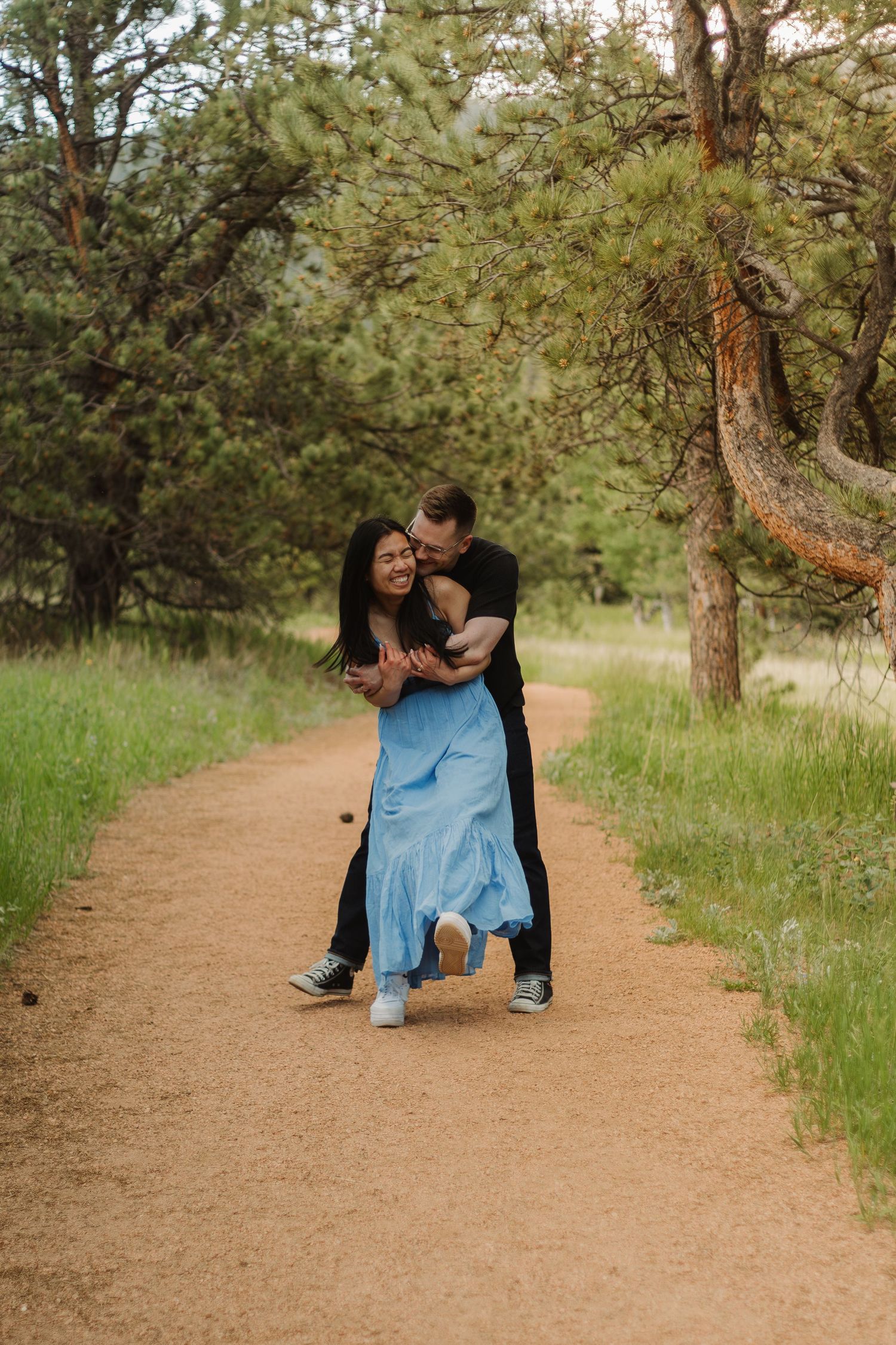 A couple embraces on a dirt path surrounded by tall pine trees and green grass.