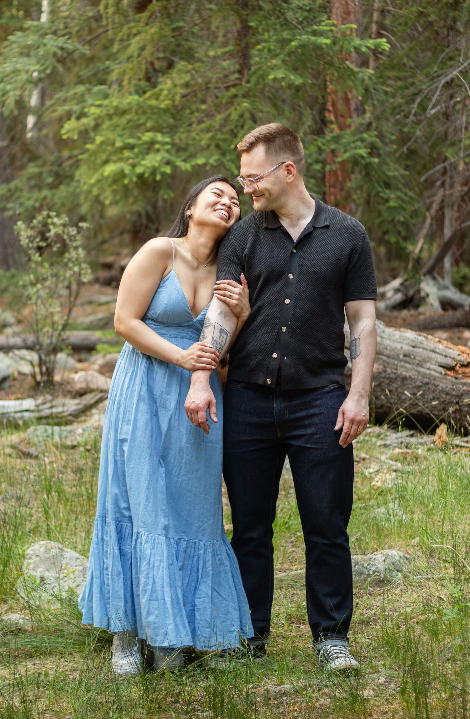 A couple stands together in a forest clearing wearing coordinated blue and black outfits.