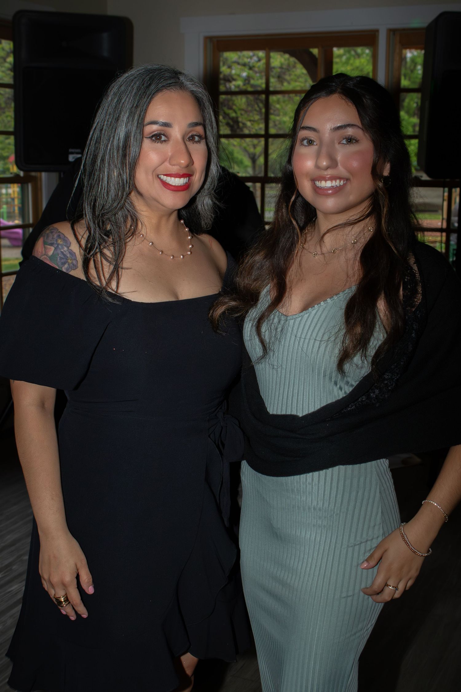 Two women pose together at an indoor event, one wearing a black dress and one in a light blue fitted dress.
