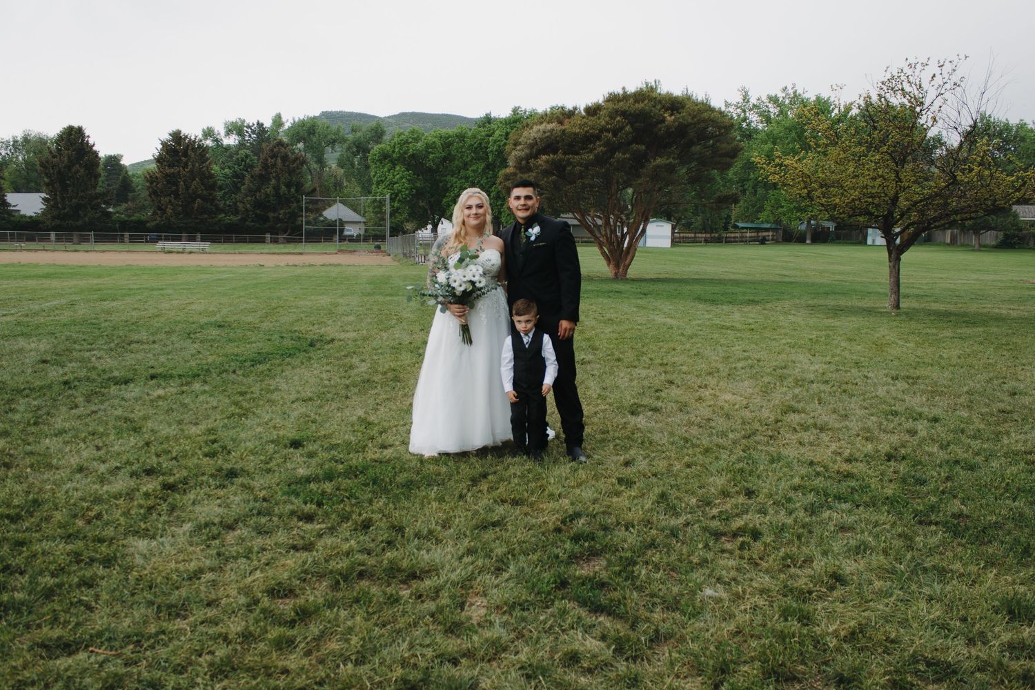Wedding party poses together on green grassy lawn with trees in background.