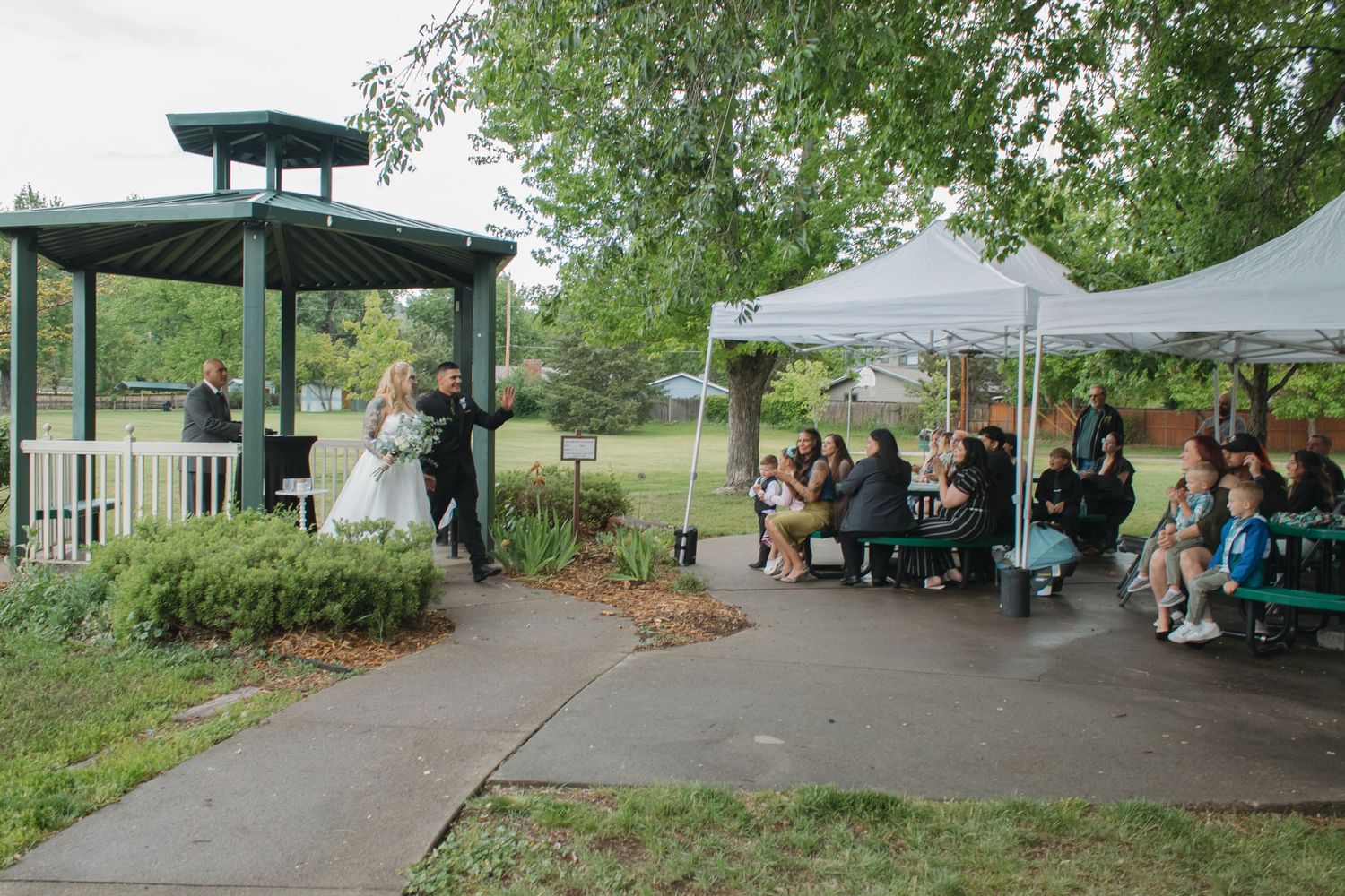 Wide view of a park pavilion wedding setup with guests gathered under white tents.