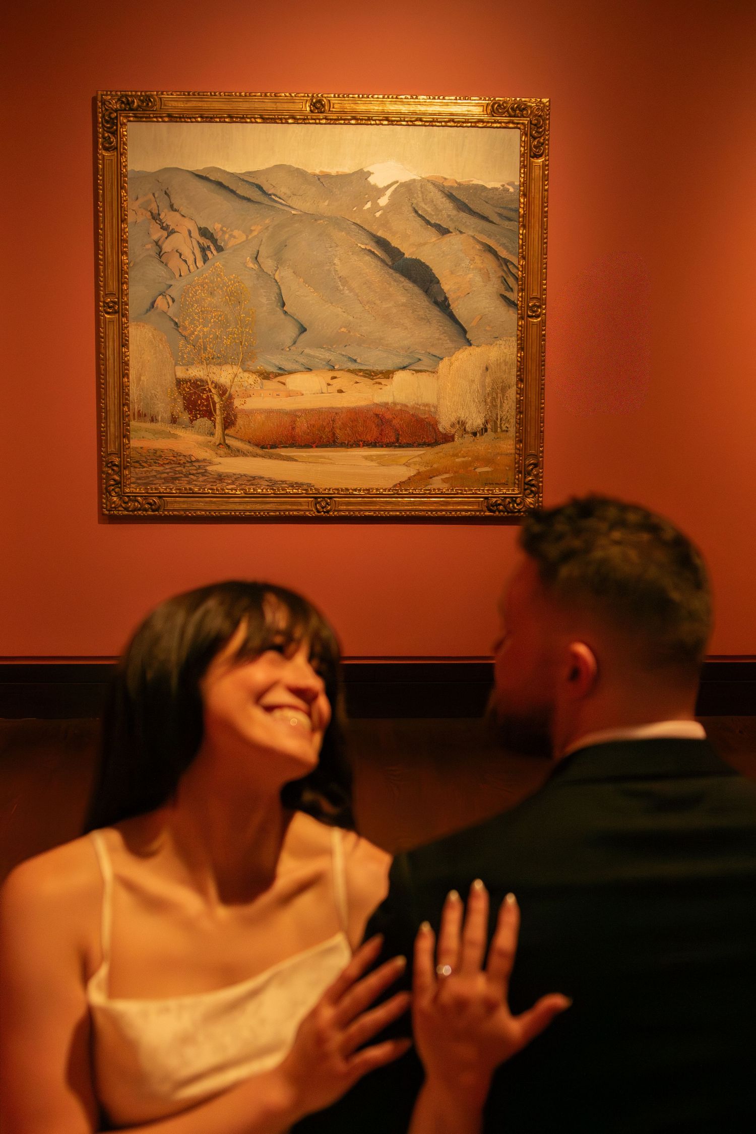 A couple shares romantic moments in front of landscape paintings in a rust-colored gallery space.