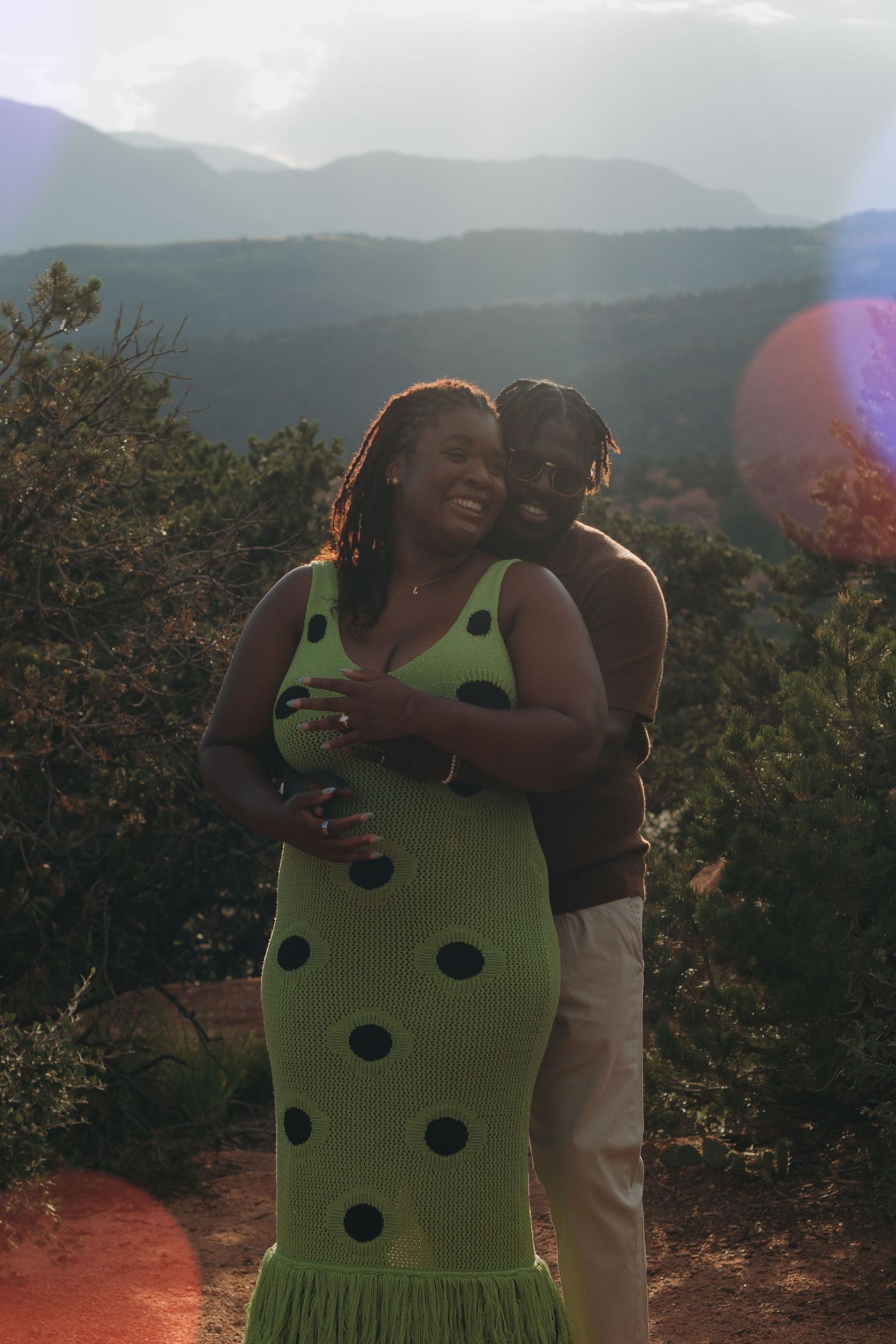 A couple shares a tender moment on a mountain overlook with sunlight creating lens flares in the foreground.