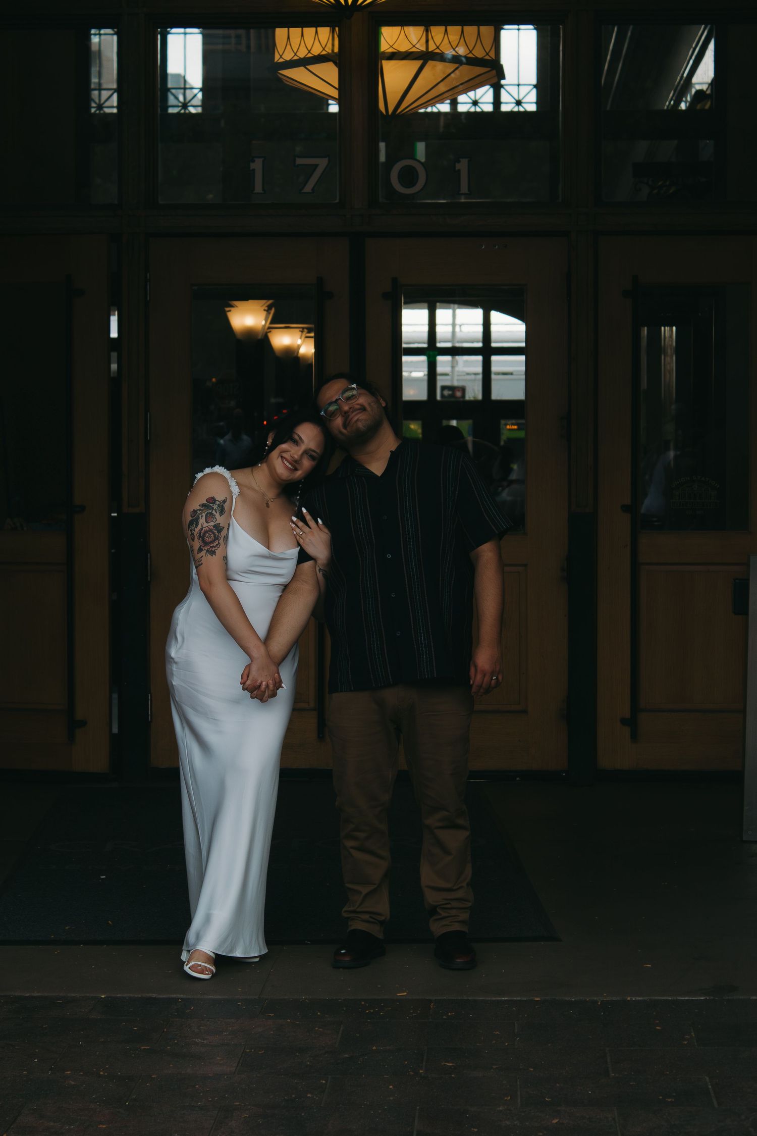 A couple in contrasting outfits poses together in a dimly lit historic building hallway.