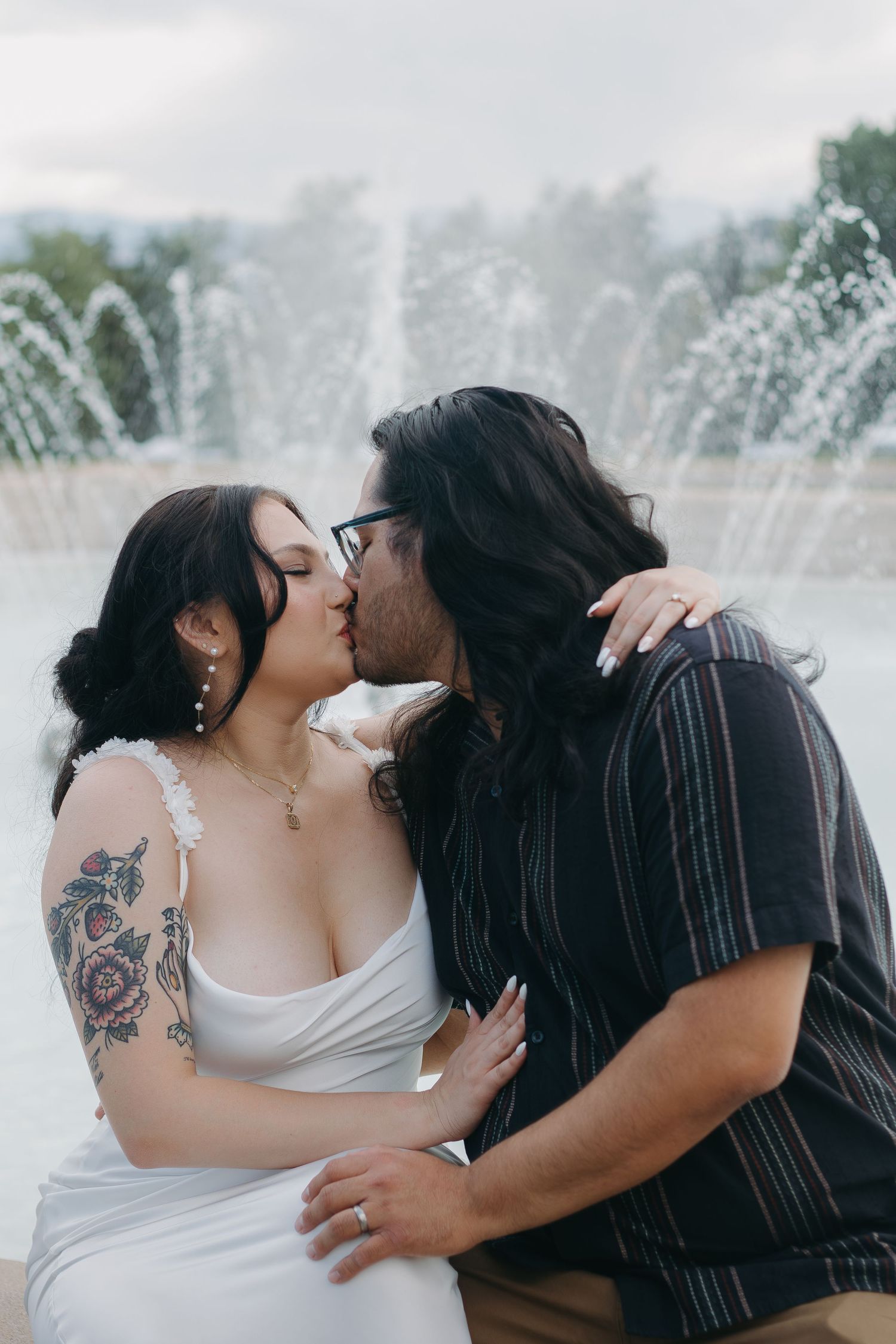 A romantic couple embraces near a flowing fountain on a cloudy day.