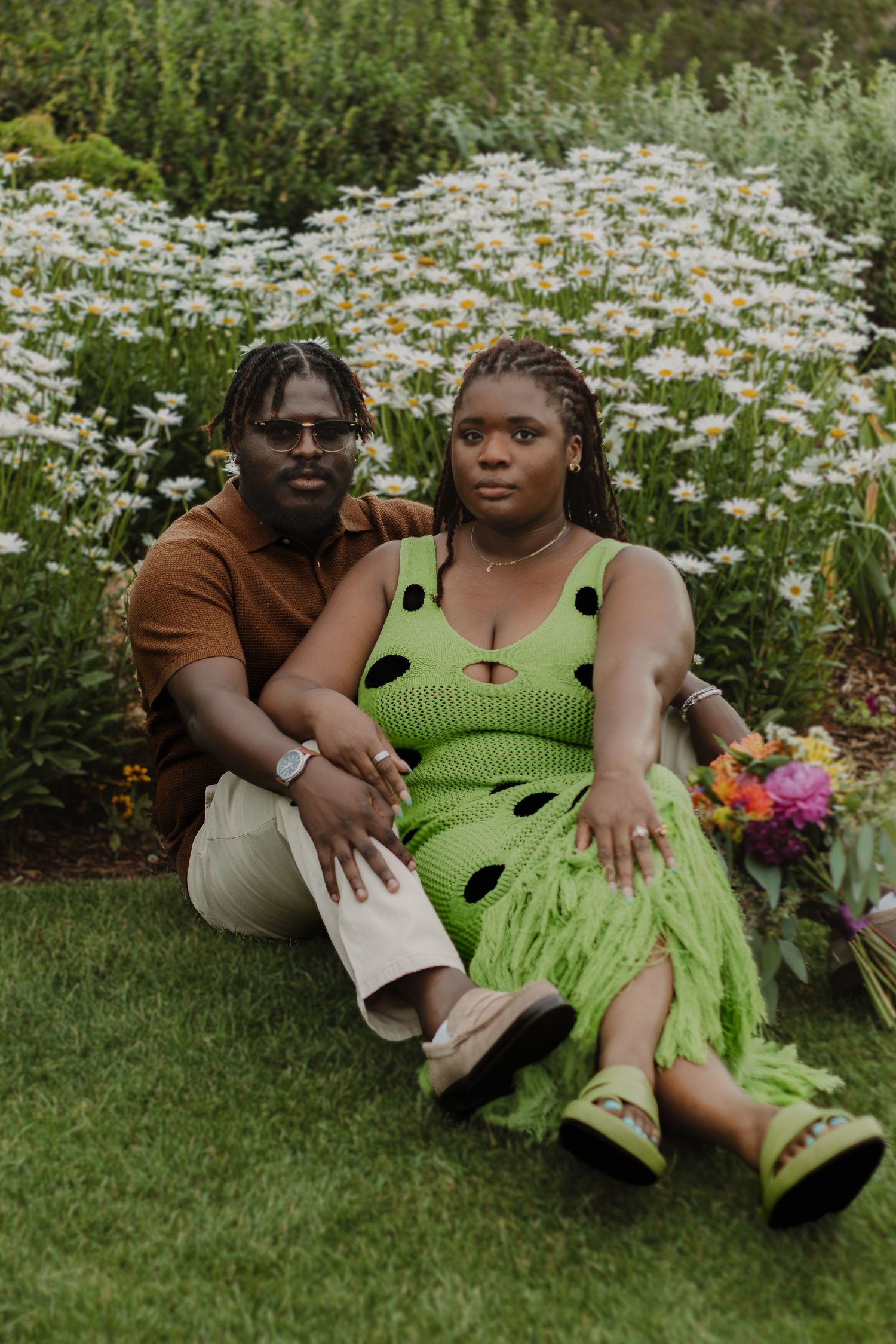 A couple relaxes on grass surrounded by blooming daisies during an outdoor photoshoot.