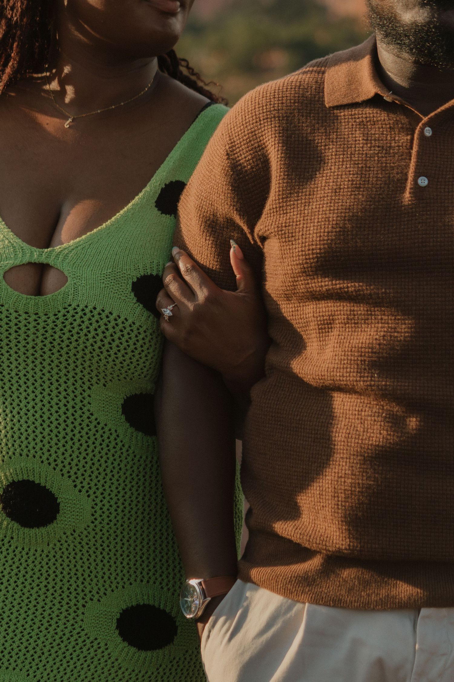 A tender embrace wearing brown and green knit sweaters against a natural outdoor backdrop.