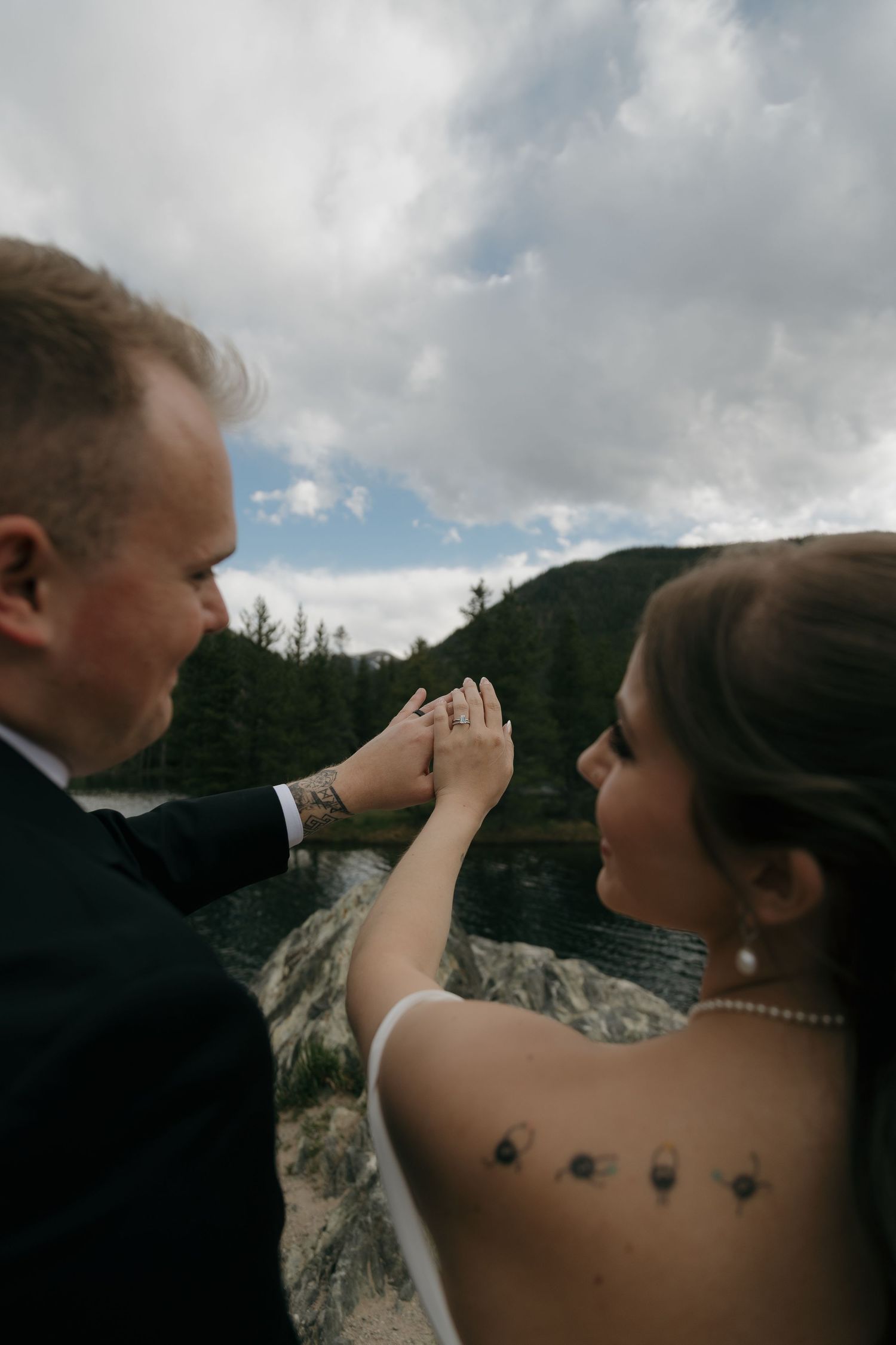 Couple admiring wedding ring together near mountain lake with dramatic cloudy sky backdrop.