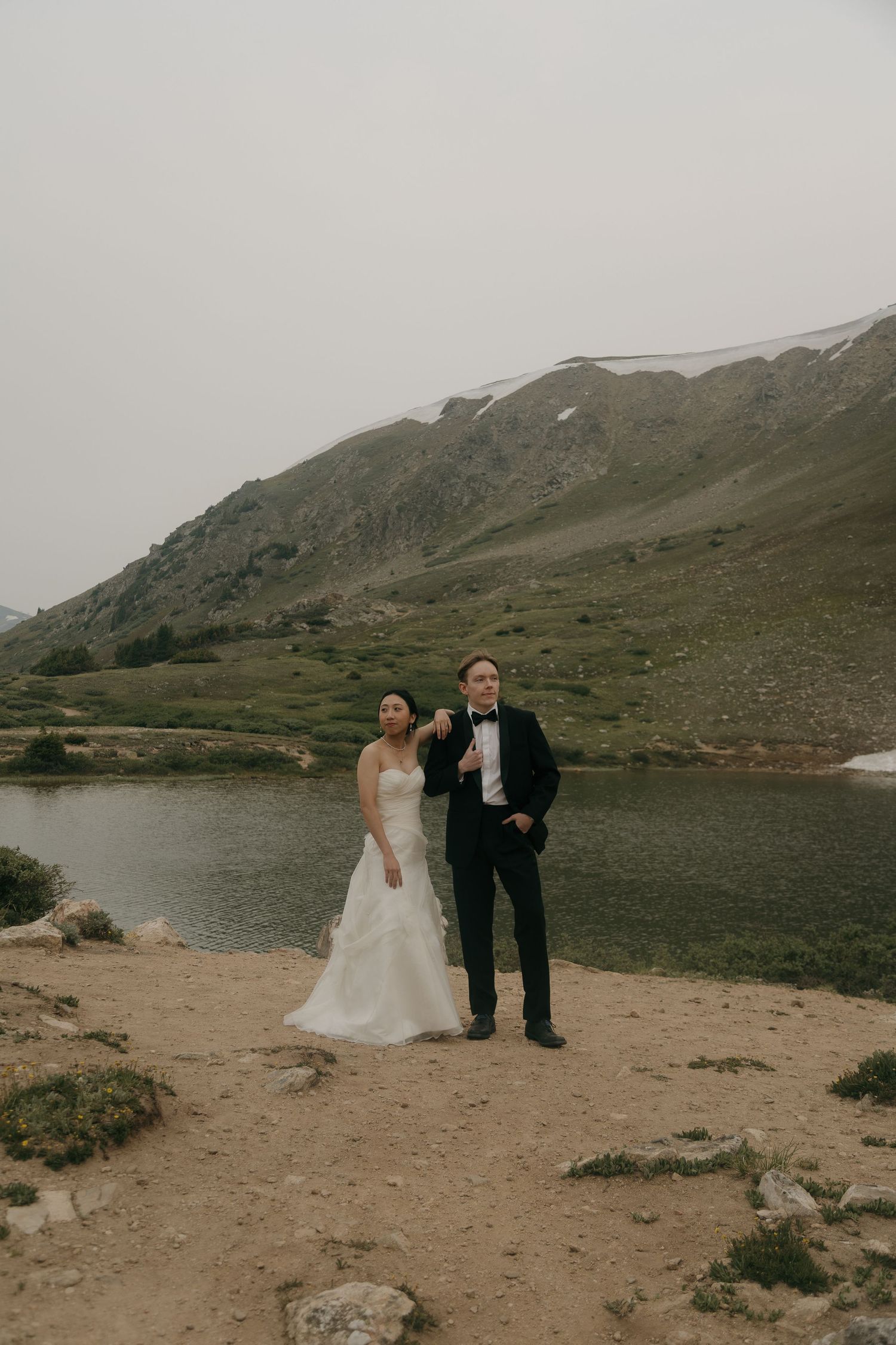 Bride and groom pose by a mountain lake, she in a white gown, he in a black tuxedo, on their elopement day.