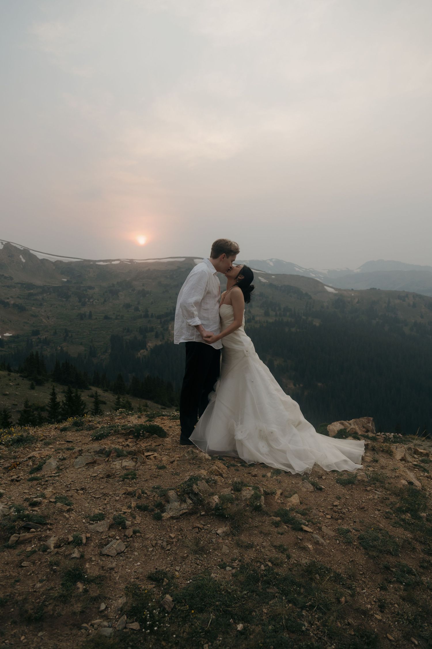 Bride and groom share a romantic kiss on a rocky mountain peak with a hazy sunset glowing behind them.