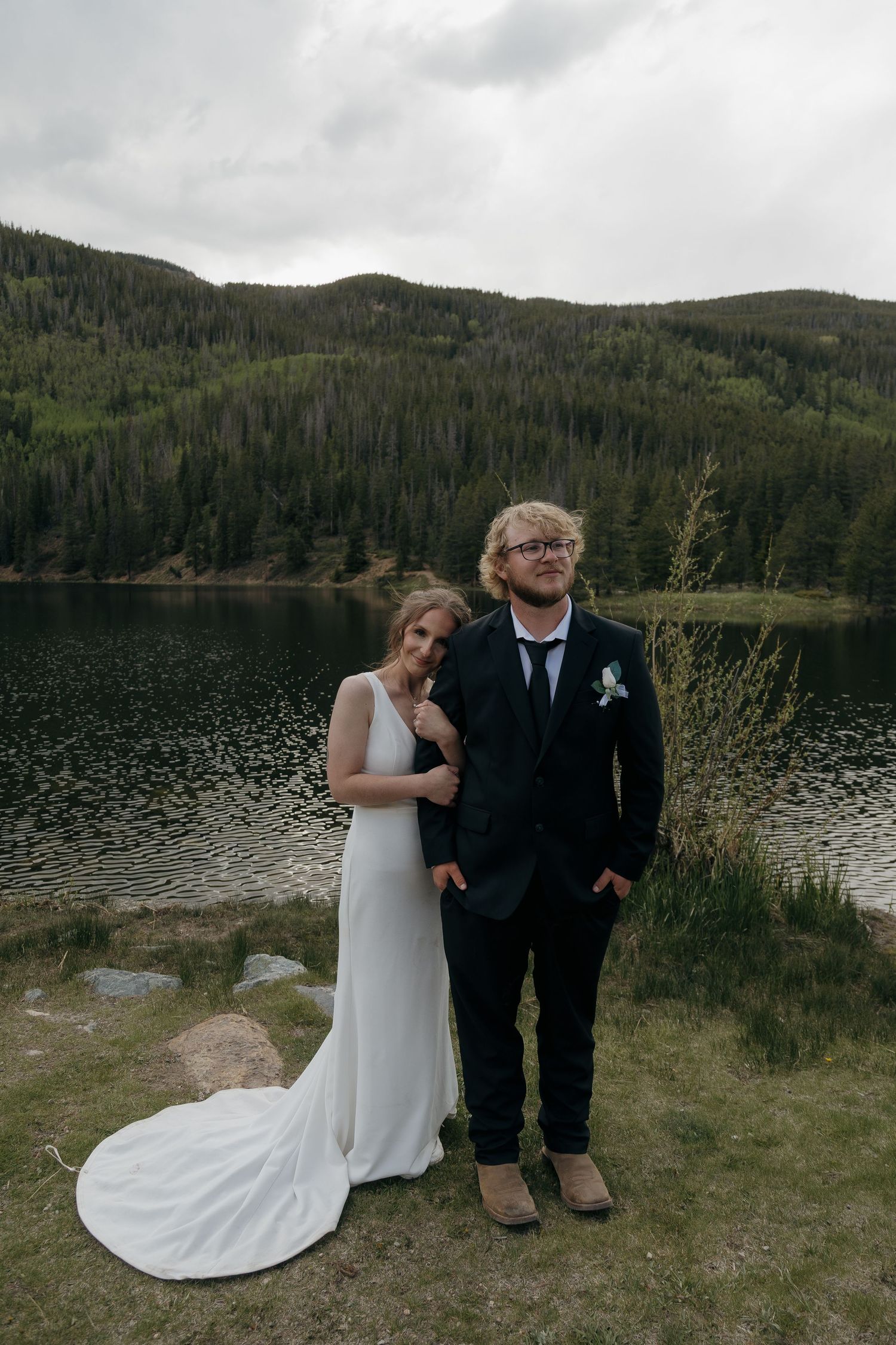 Bride and groom pose together before a serene mountain lake surrounded by pine forests.