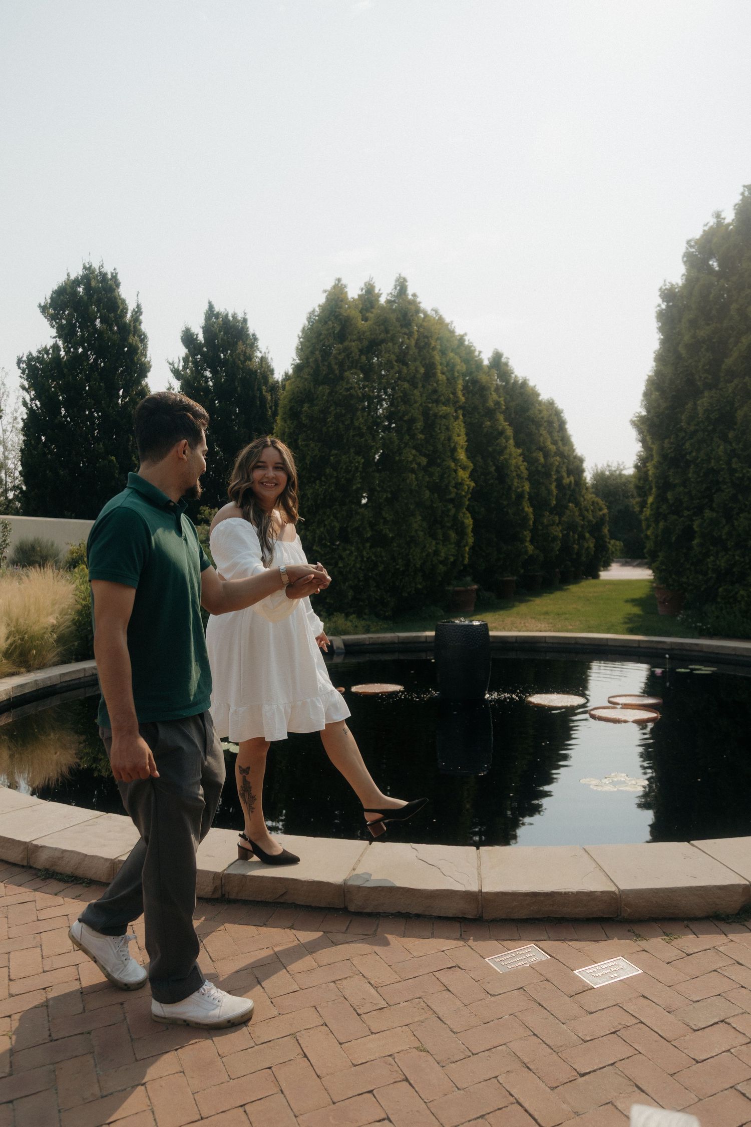 Couple walking and laughing together beside a circular reflecting pool surrounded by tall evergreen trees.