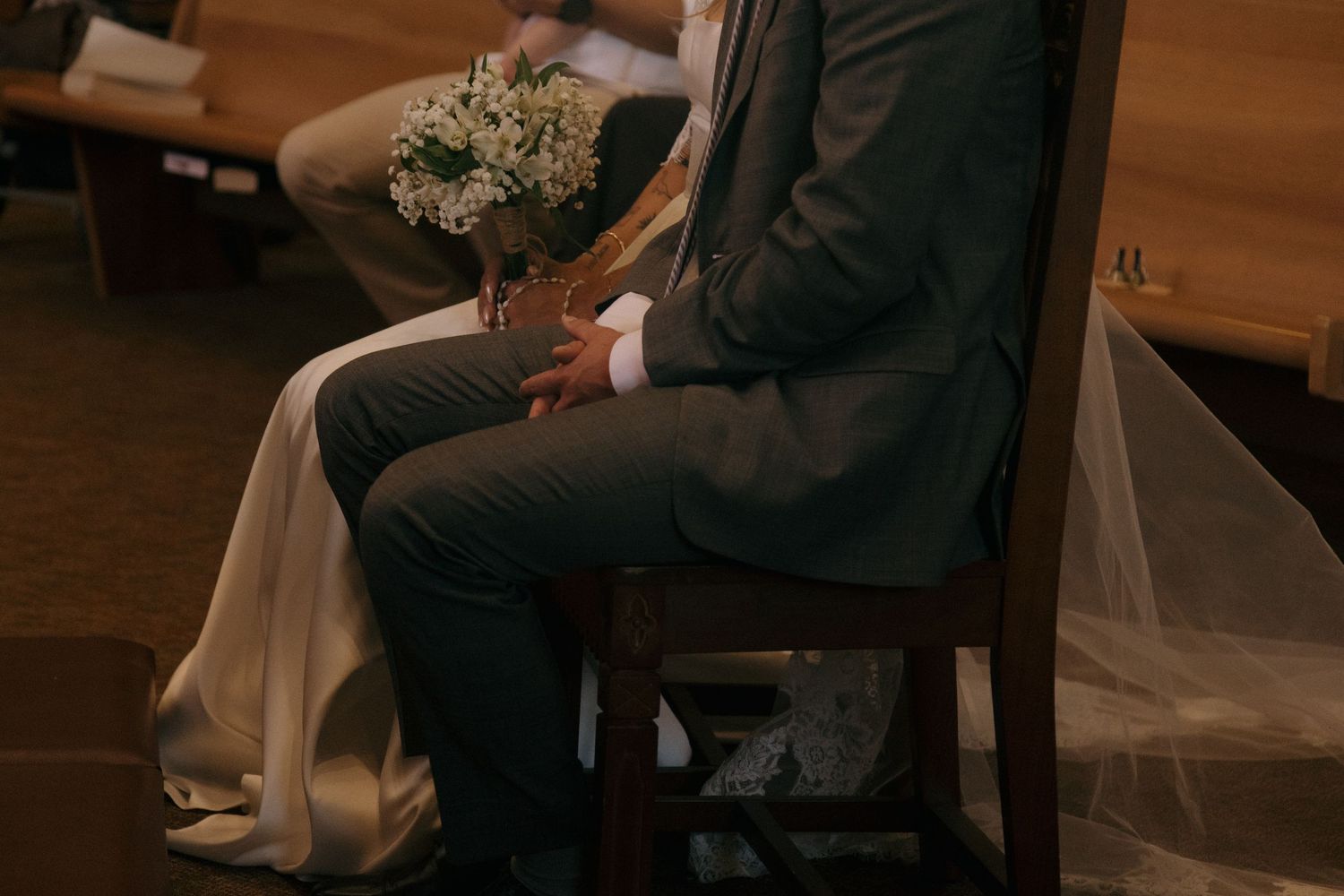 Close-up of bride and groom seated in church chairs, bride holding baby's breath bouquet.