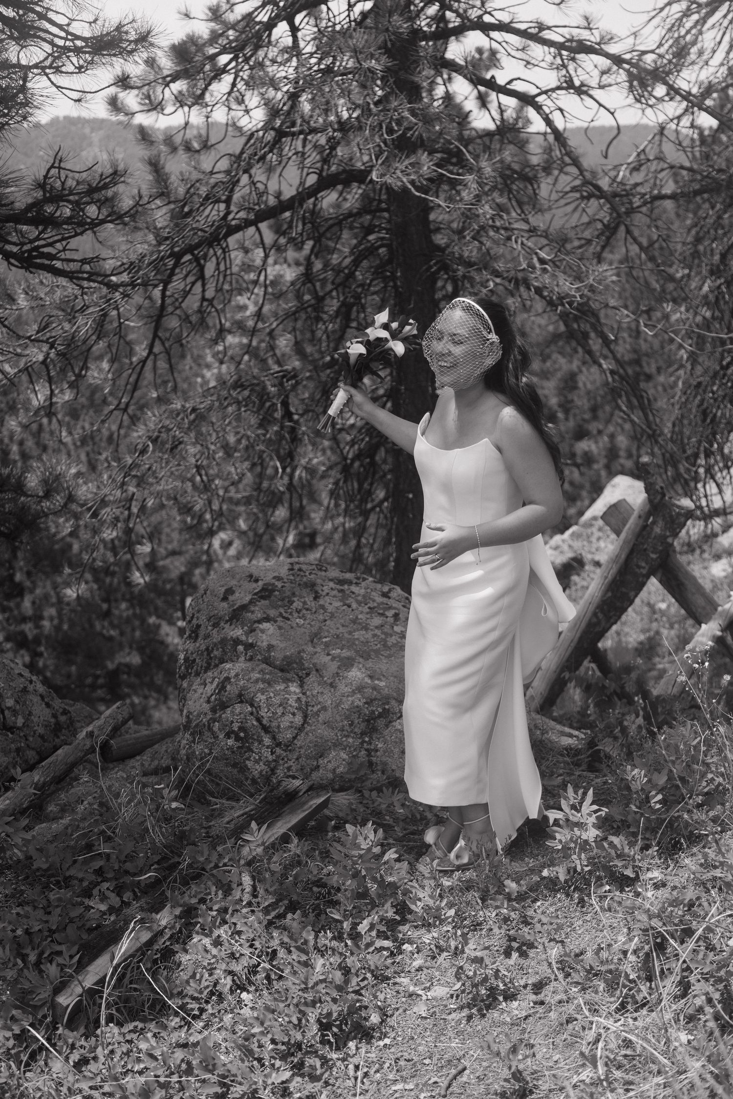 Black and white photo of smiling bride in white dress holding bouquet among pine trees in natural outdoor setting.