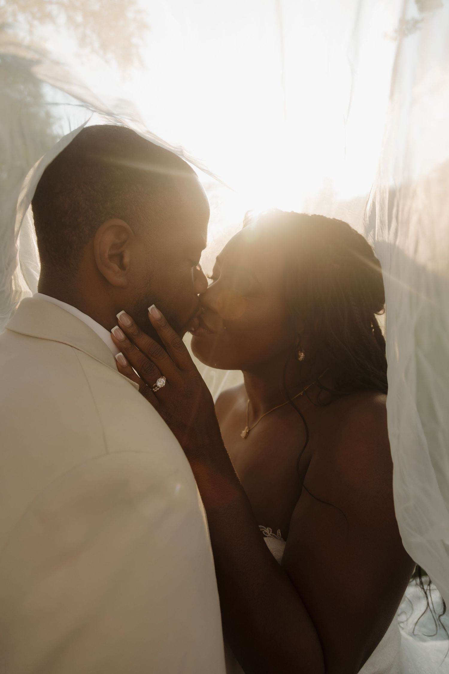 Bride and groom share an intimate kiss under a veil with warm golden backlight glowing around them.