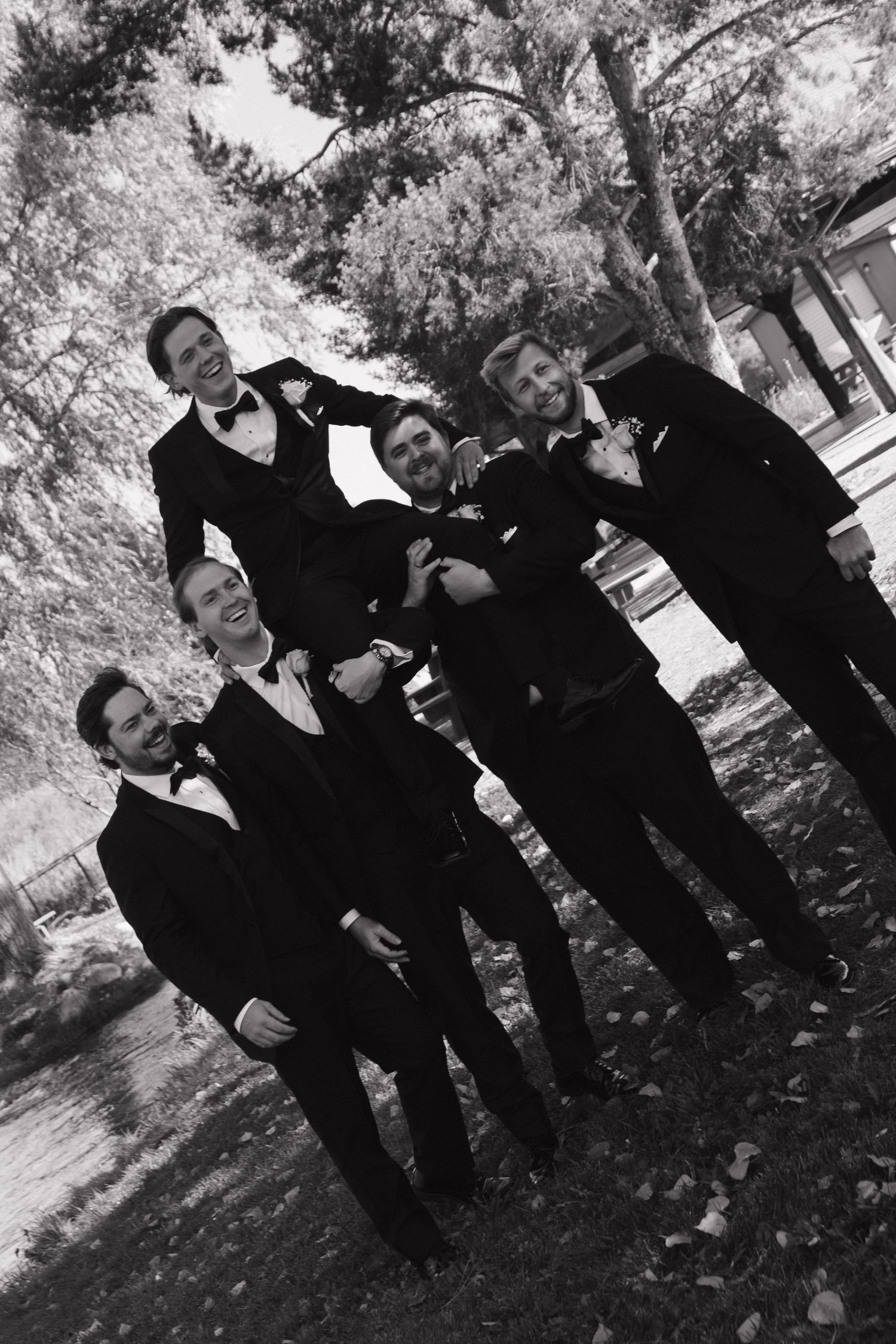 Wedding party in black formal attire poses together outdoors in a candid group shot.