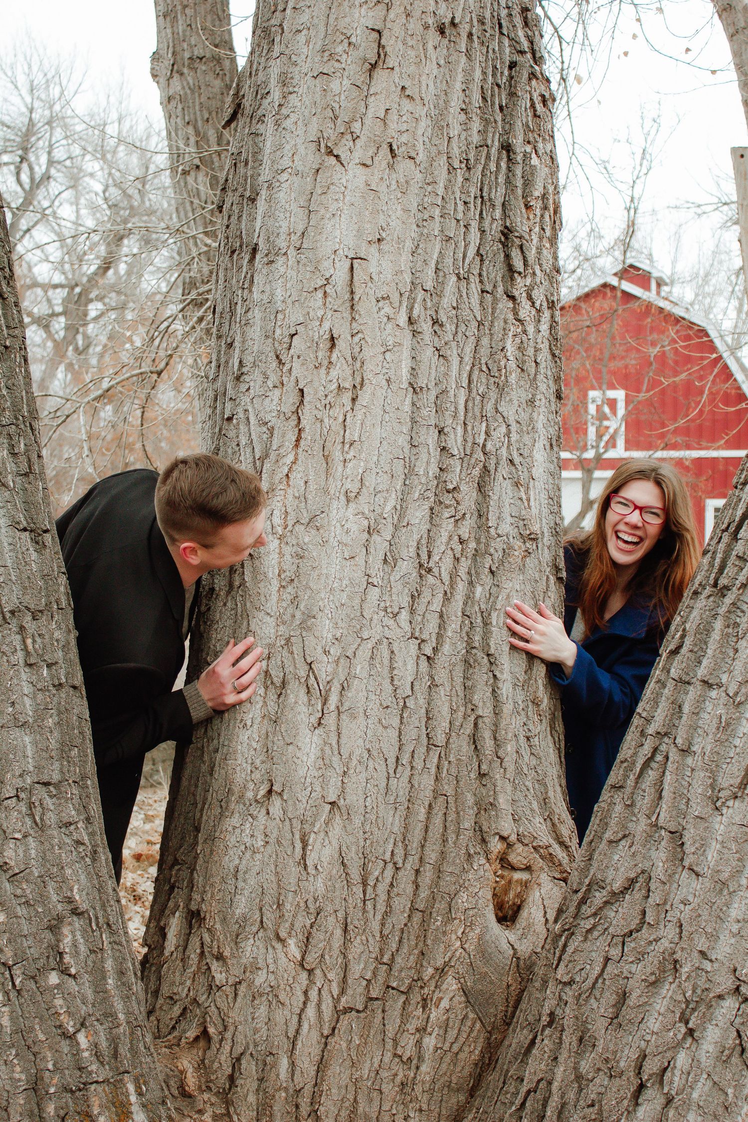 A playful couple peeks around opposite sides of a large tree trunk with a red barn in background.
