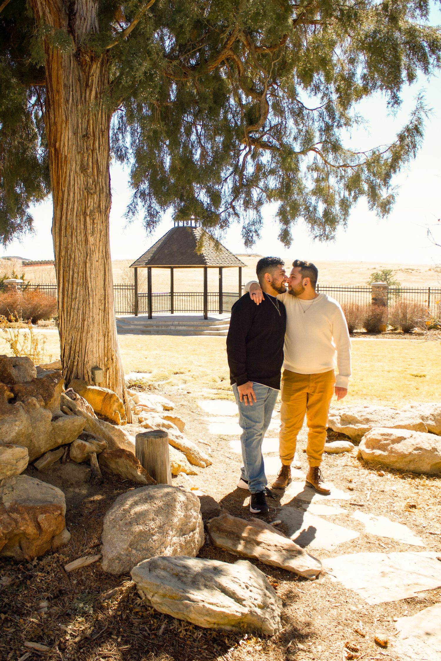 Two people stand together by a gazebo and large tree in a desert landscape.