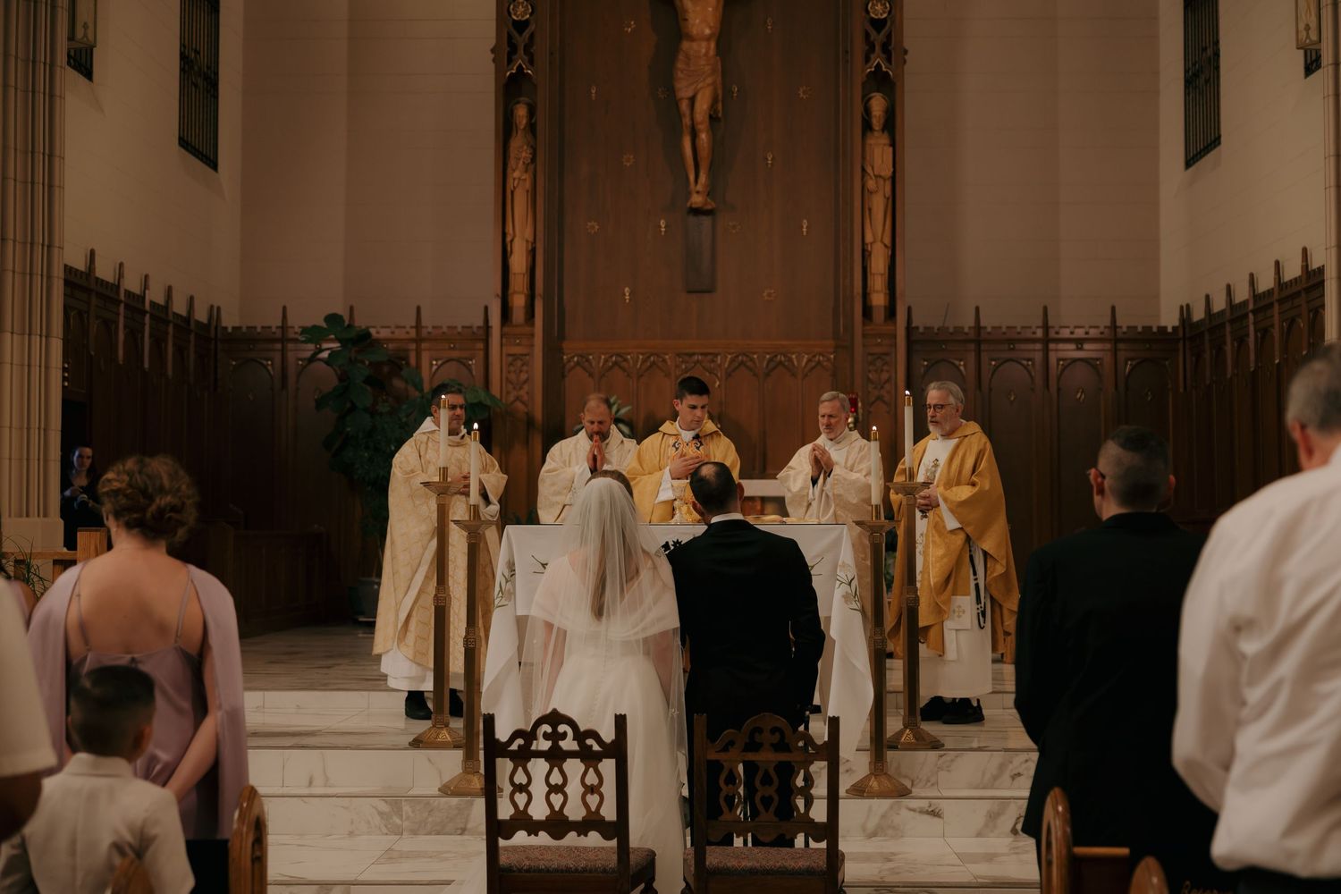 A bride and groom kneel before clergy at a Catholic church wedding ceremony altar with golden vestments.