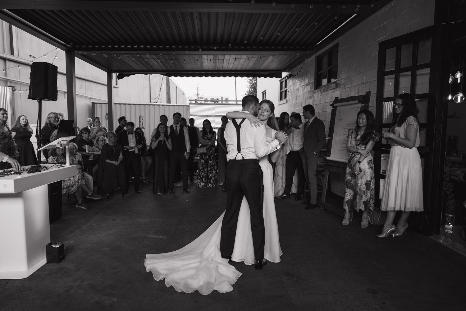 Black and white photo of bride and groom sharing first dance surrounded by wedding guests at reception.