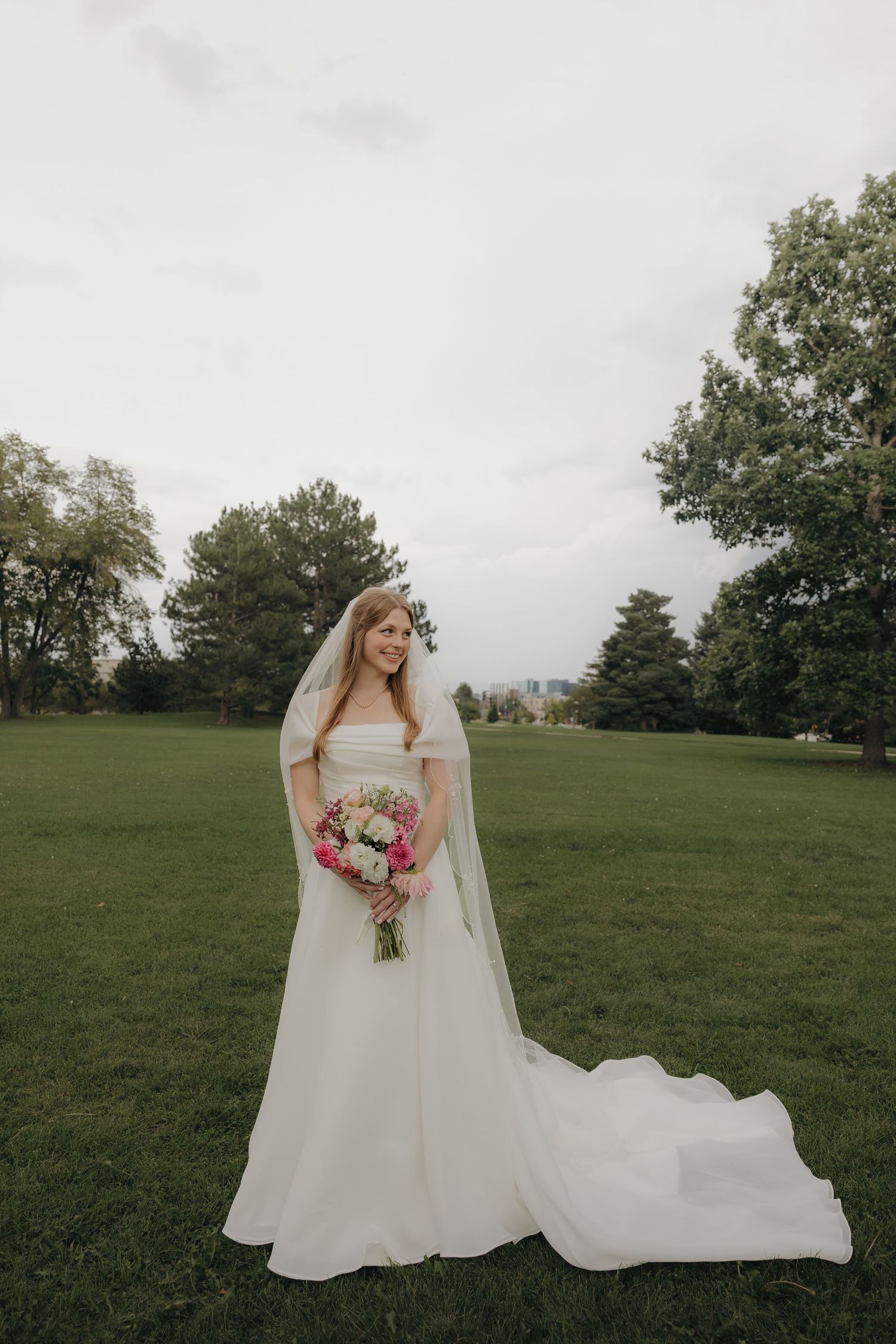 Elegant bride in off-shoulder white gown with cathedral veil holds pink bouquet in lush green park.