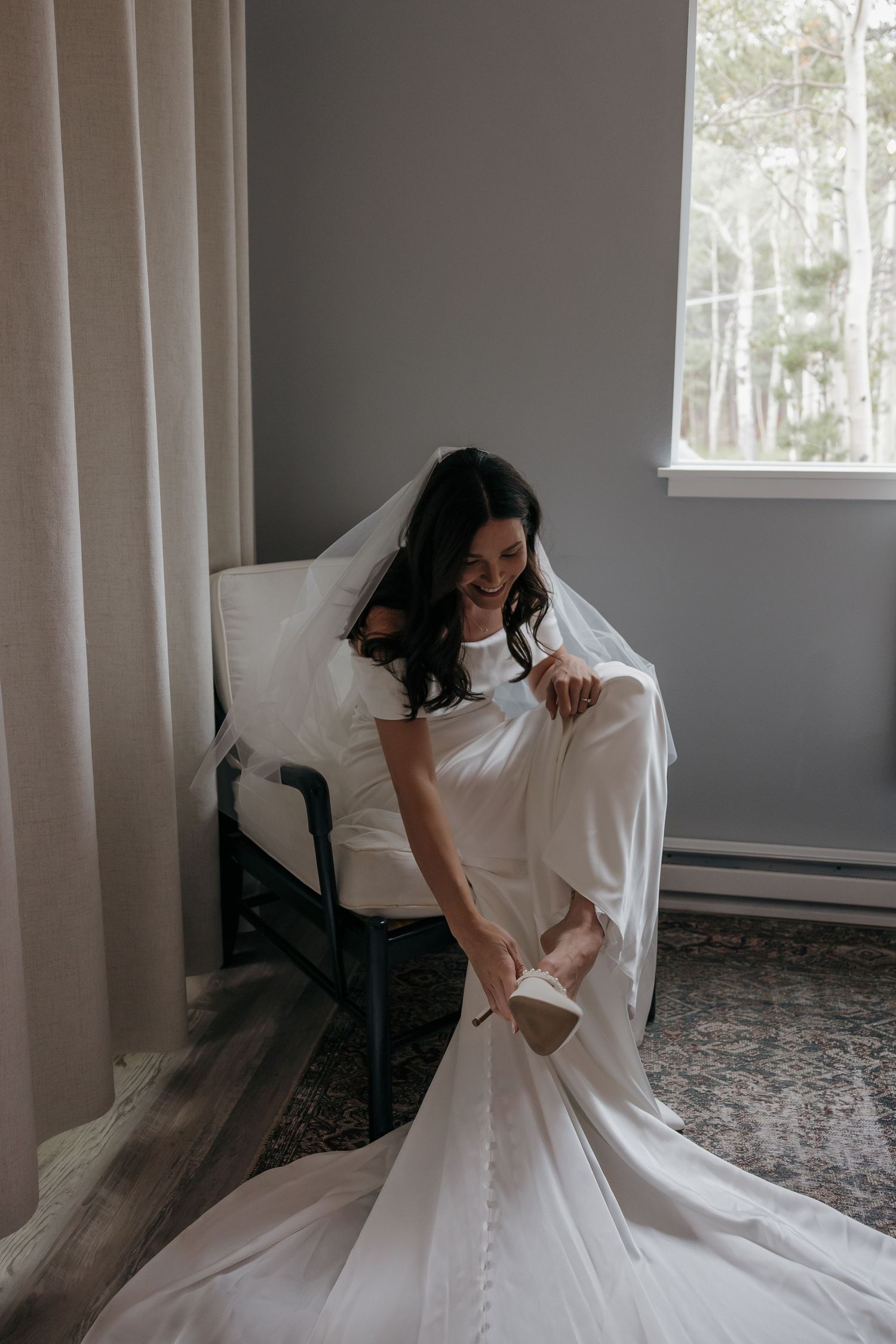 Bride in off-shoulder white gown and veil sits in chair putting on heels near window.